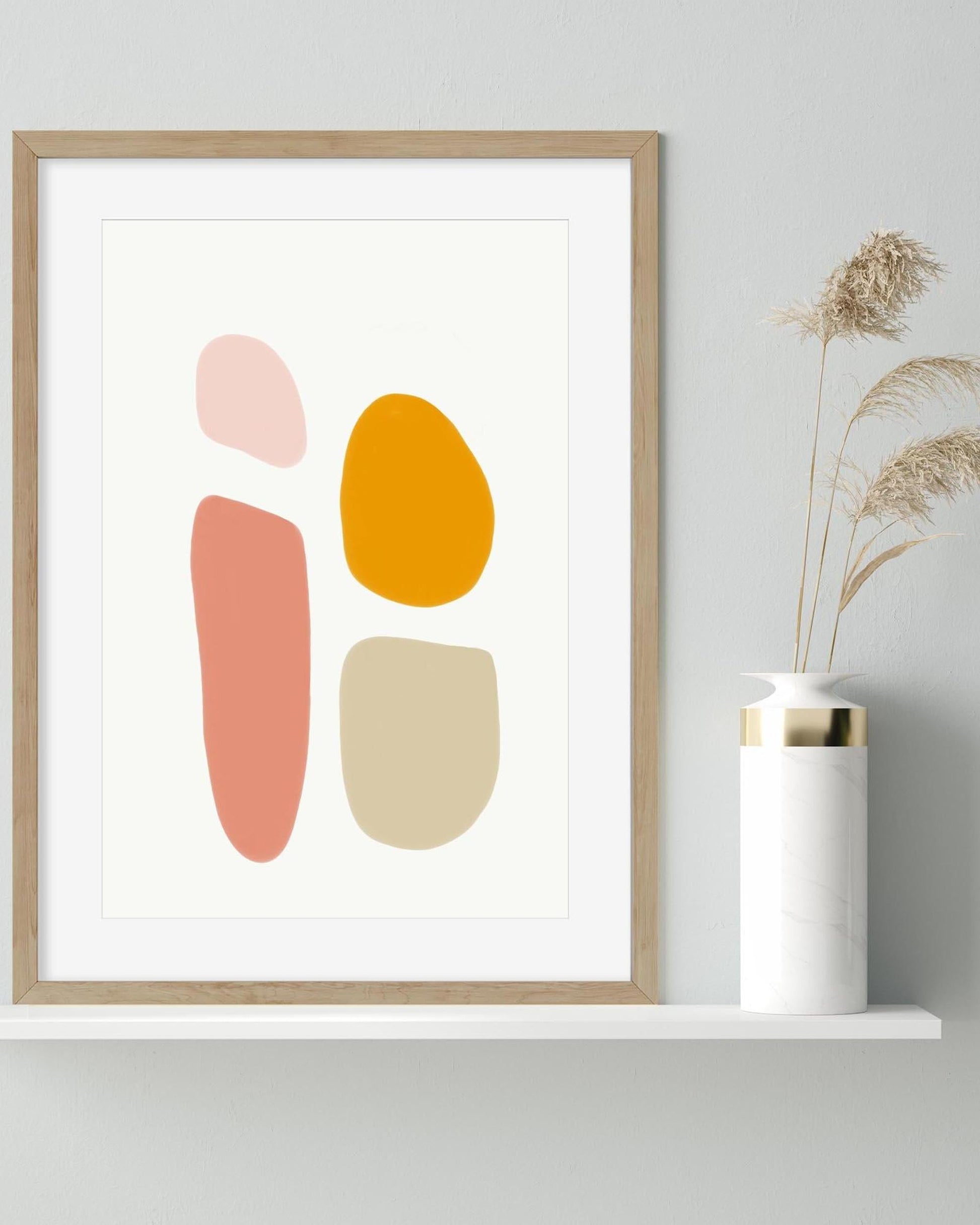 Framed abstract art print with colorful shapes on a white wall, next to a vase with dried plants.