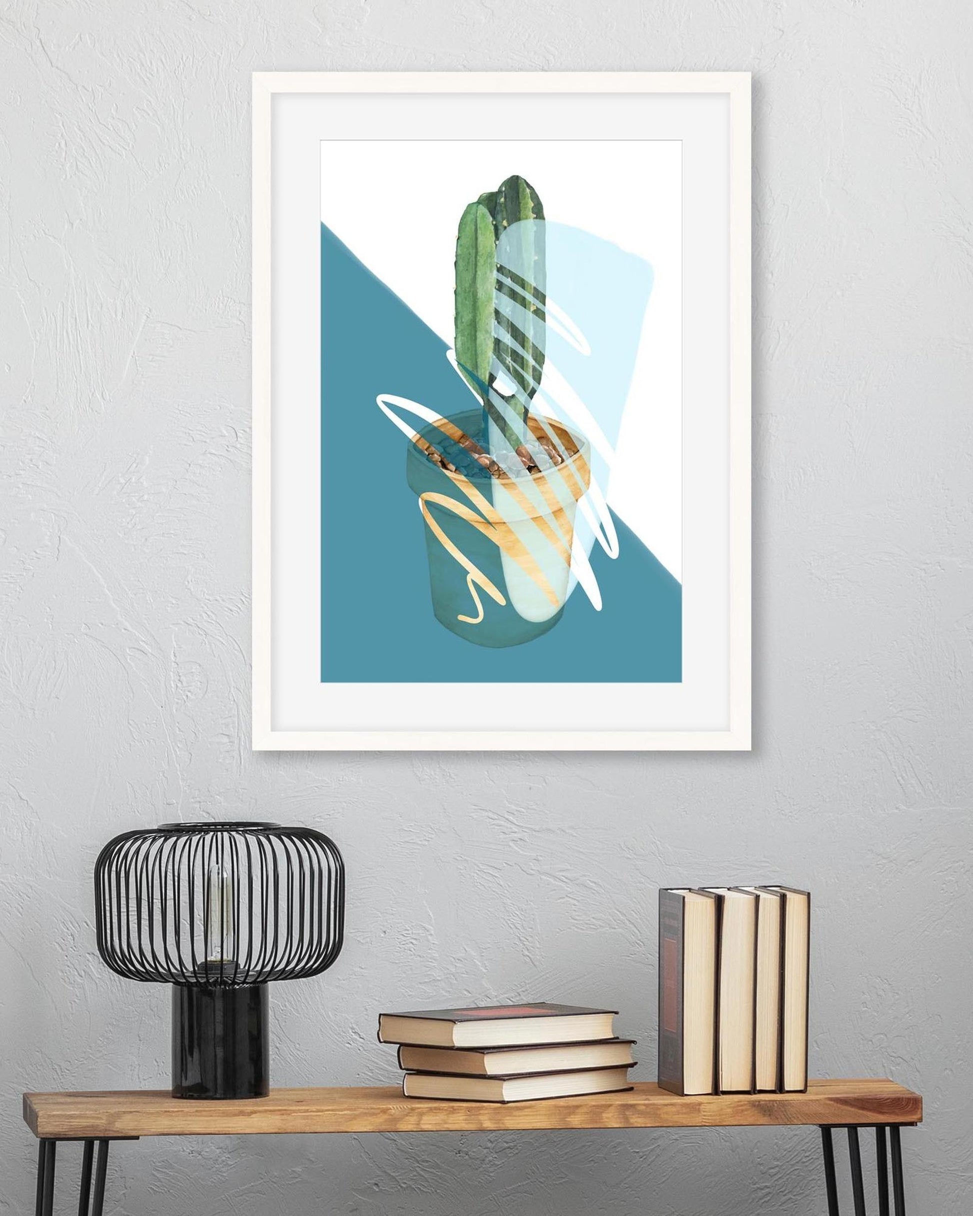 Framed abstract art print of a cactus on a wall above a wooden shelf with books and a lamp.