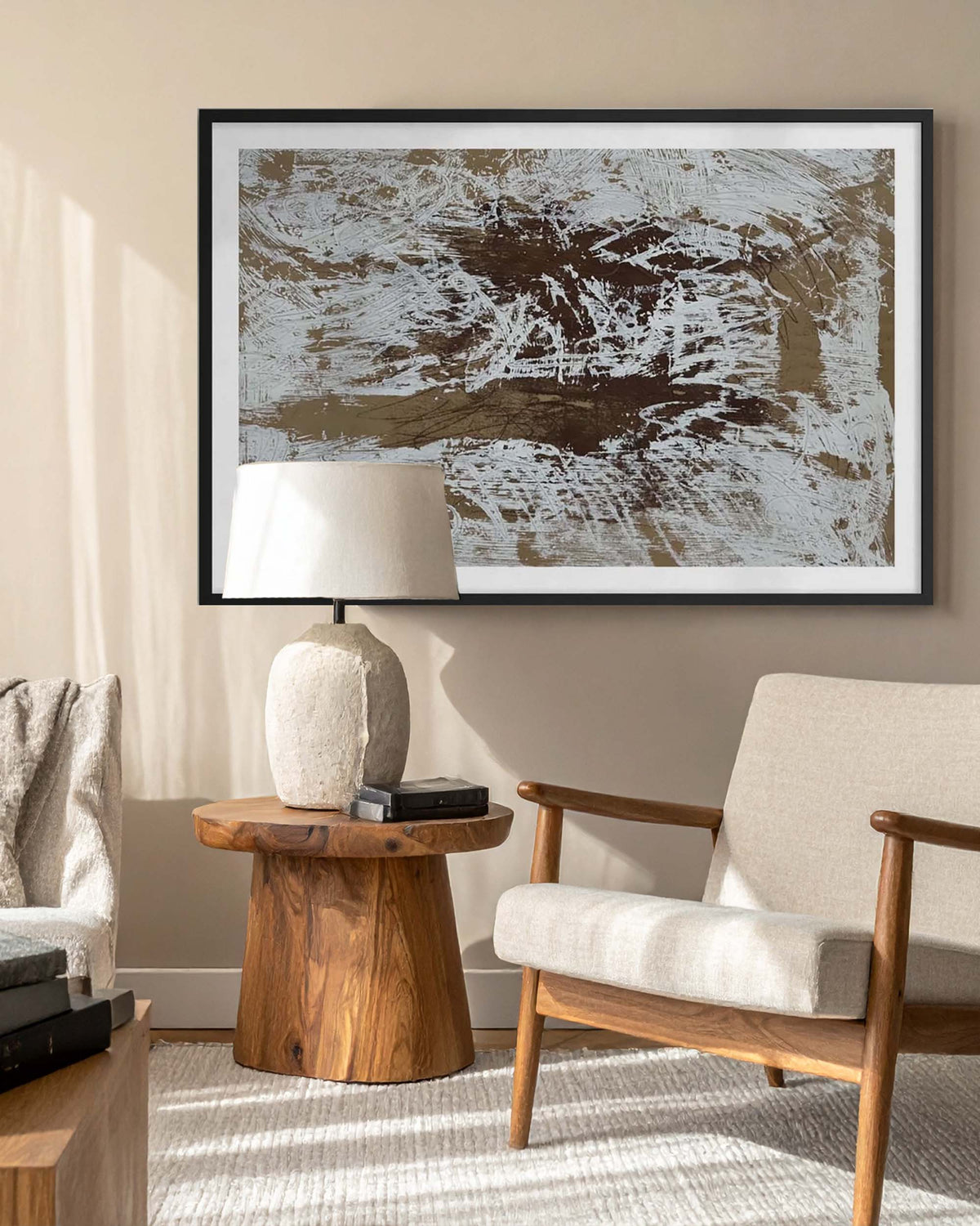 A framed original abstract painting with brown, beige and  white brushstrokes hangs on a beige wall in a modern living room setting with a wooden side table, lamp, and armchair.