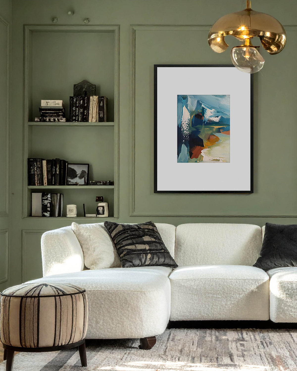A framed limited edition abstract art print with a white mat and black frame hangs on a sage green paneled wall above a white boucle sectional sofa in a styled living room.