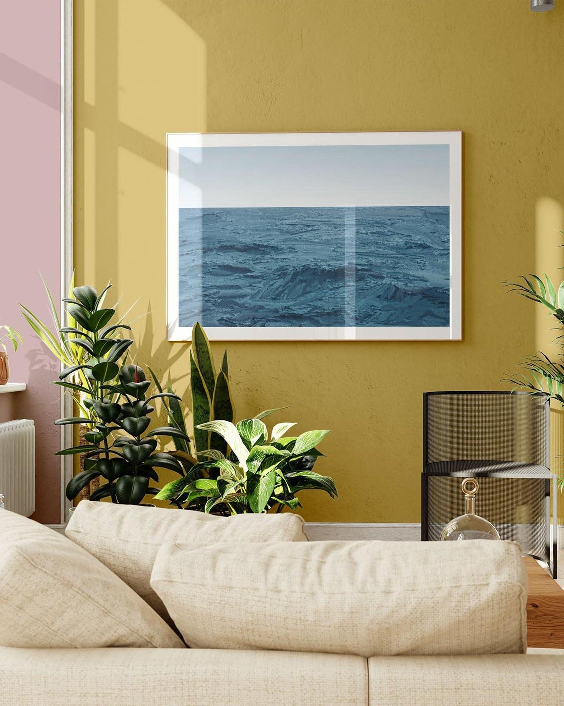 Living room with a beige sofa, plants, and a framed picture of the ocean on a yellow wall.