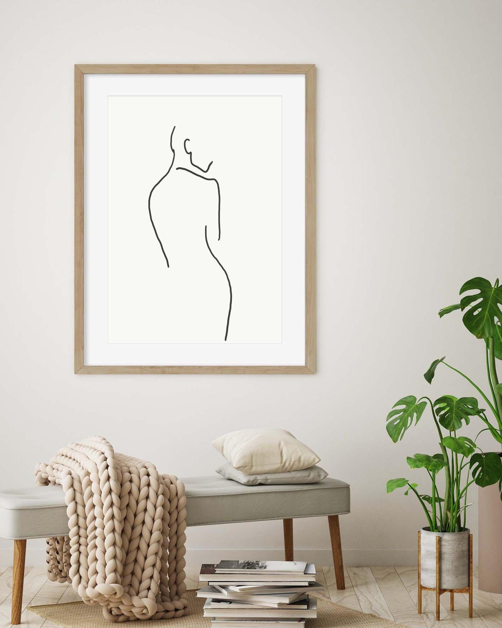 Framed line art of a woman's silhouette on a wall with a bench, books, and plant in the foreground.