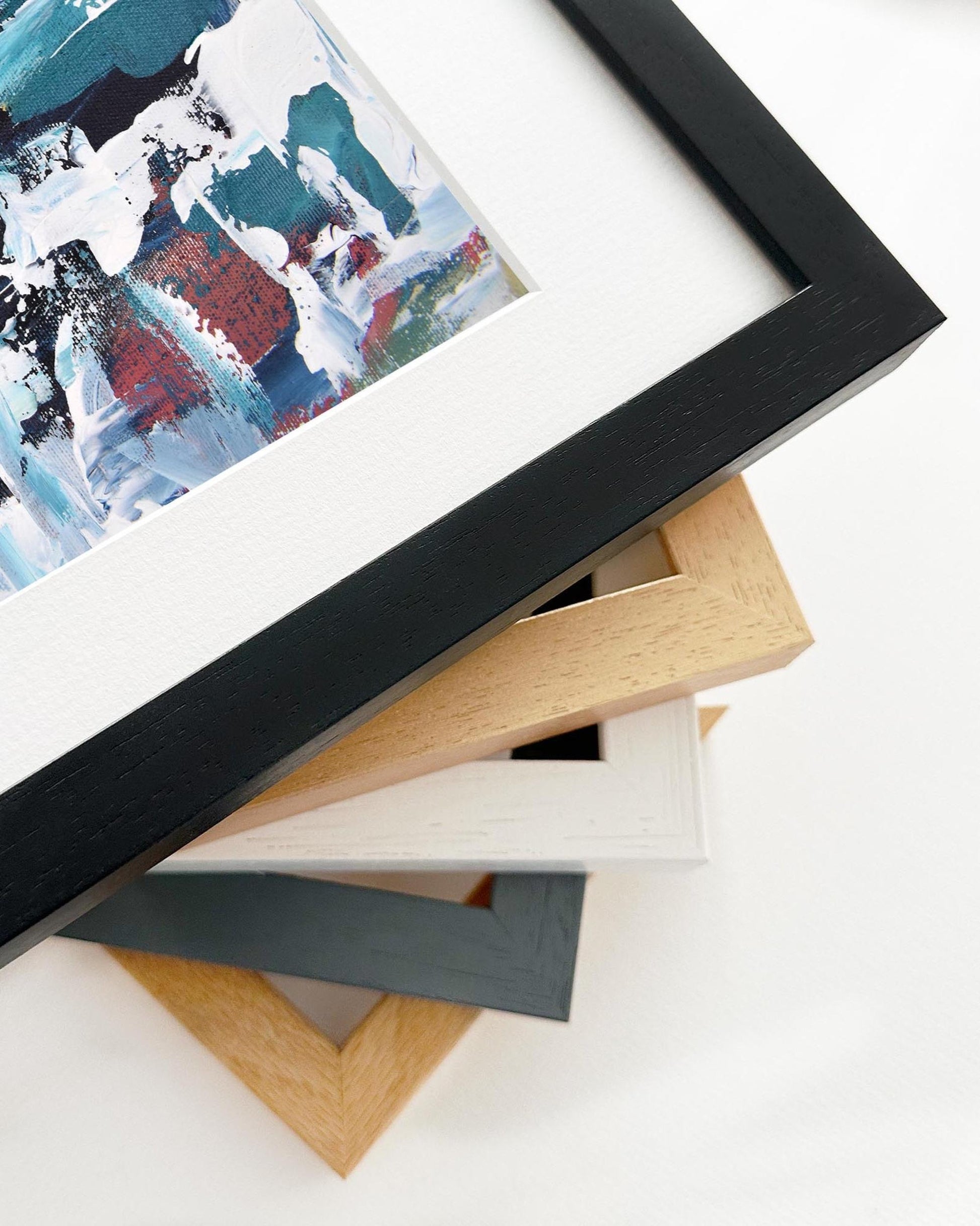 Stack of variously colored picture frames on a white surface with a framed abstract painting.