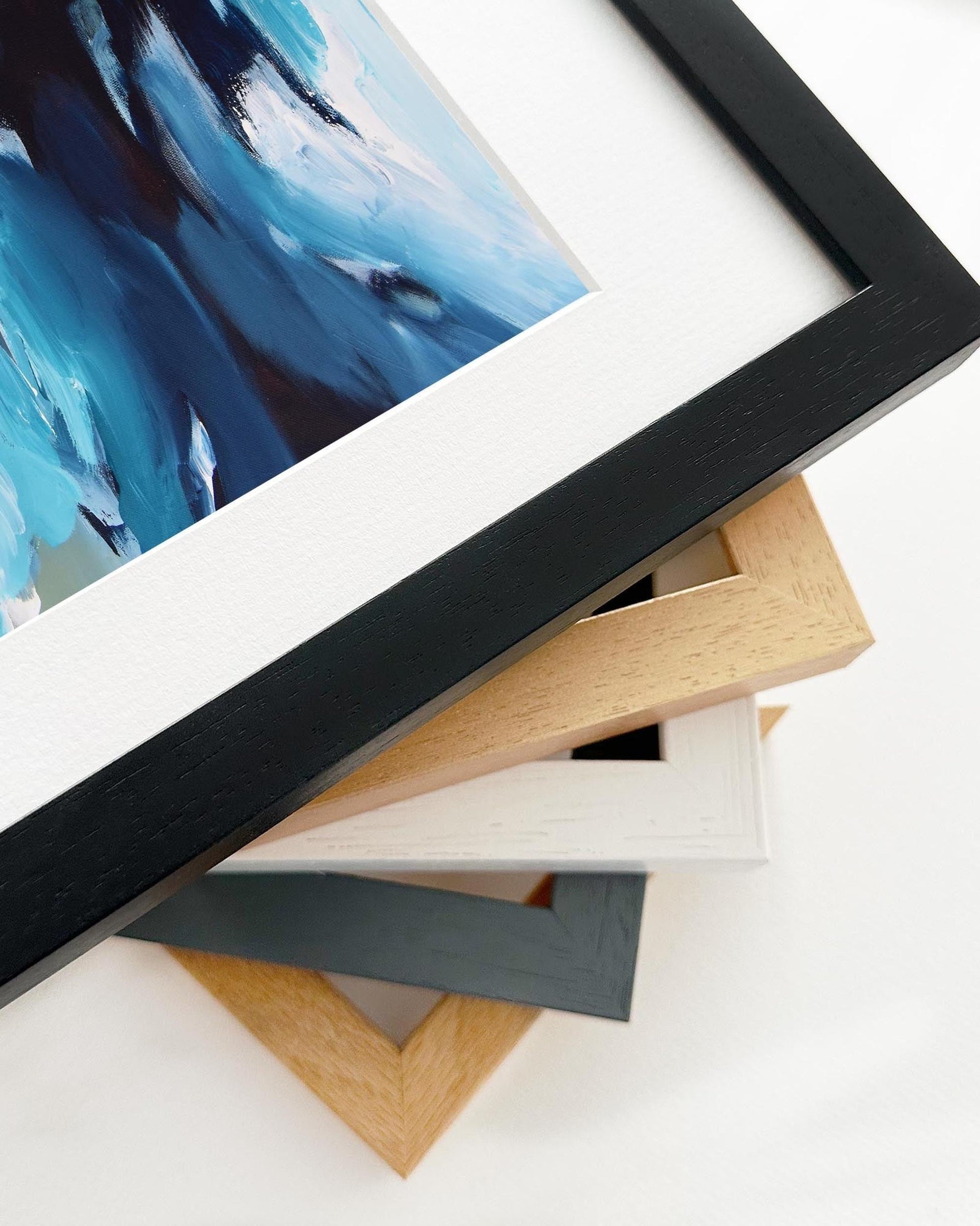 Stack of variously colored picture frames on a white surface with a blue abstract painting in the foreground.