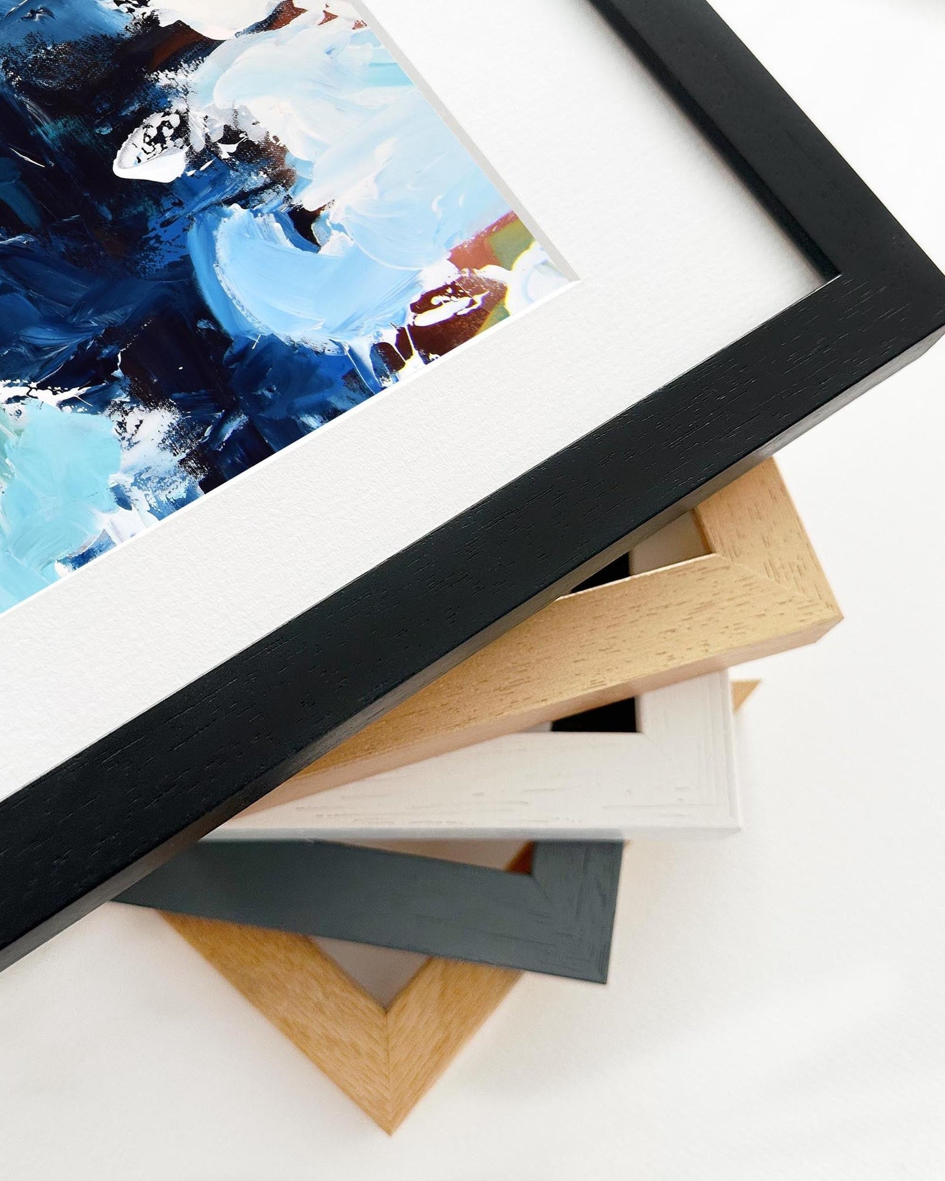 Stack of variously colored picture frames on a white surface with a framed abstract painting in the foreground.