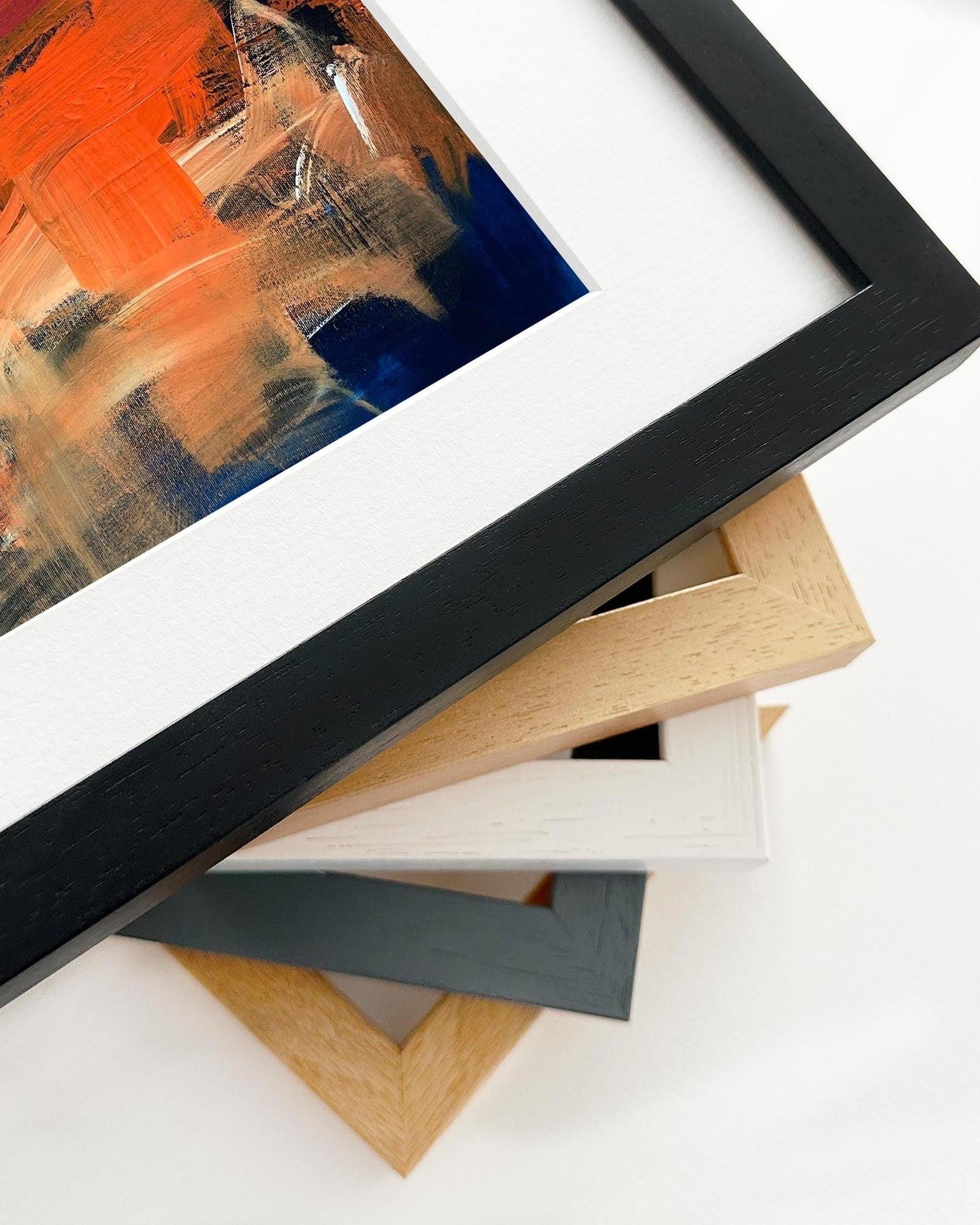 Stack of various picture frames on a white surface with a framed abstract painting.