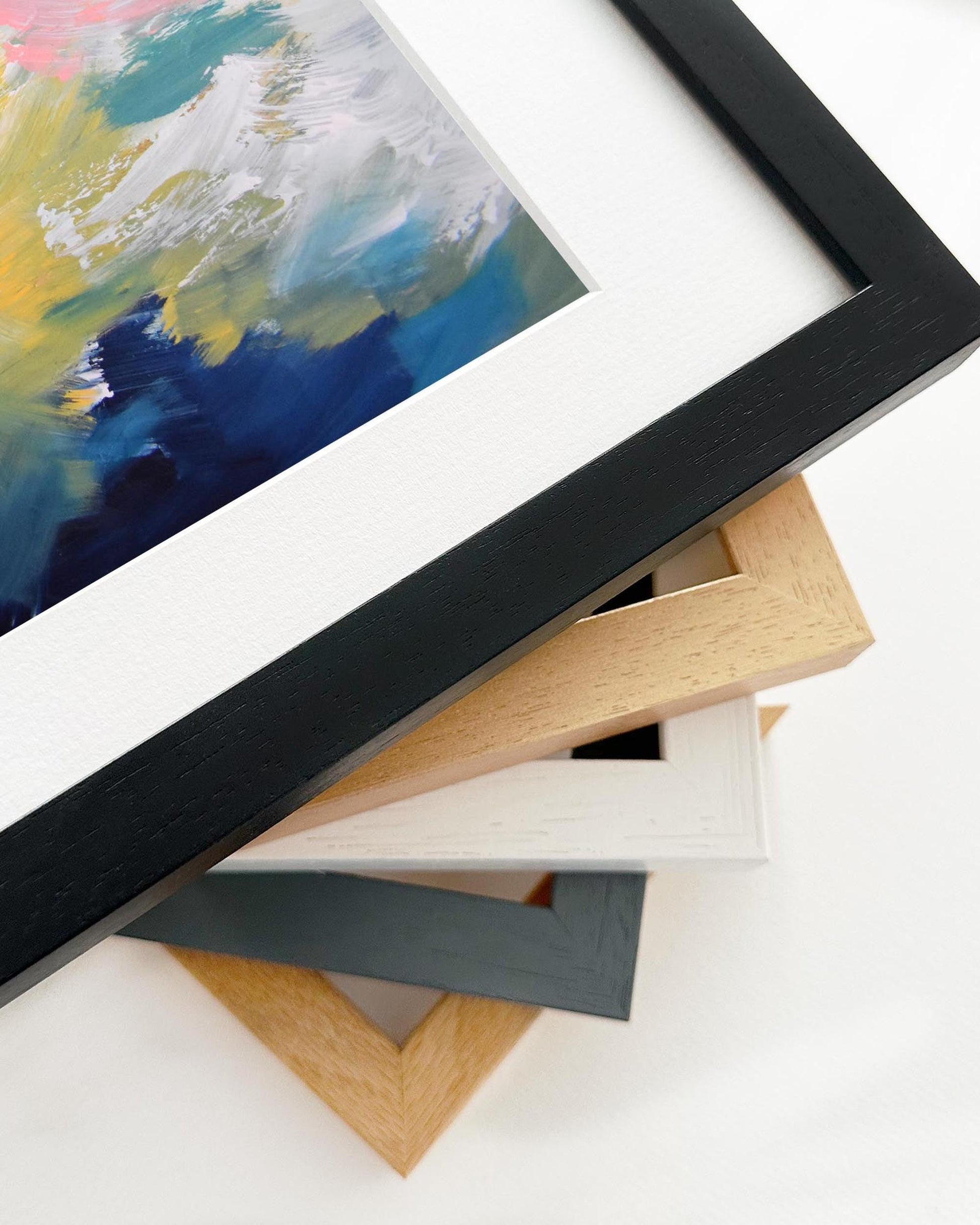 Stack of variously colored picture frames on a white surface with a colorful painting in the background.