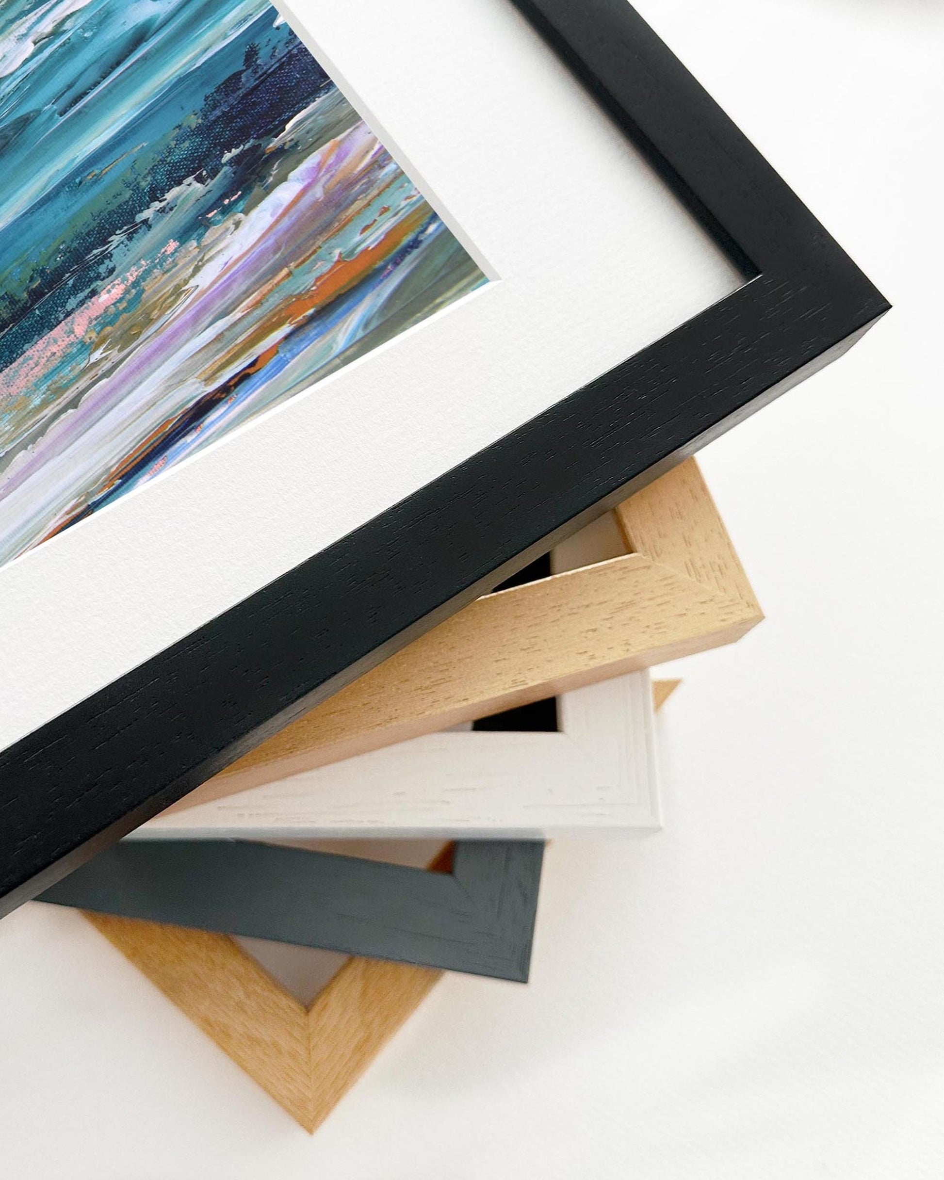 Stack of variously colored picture frames on a white surface with a framed artwork in the foreground.