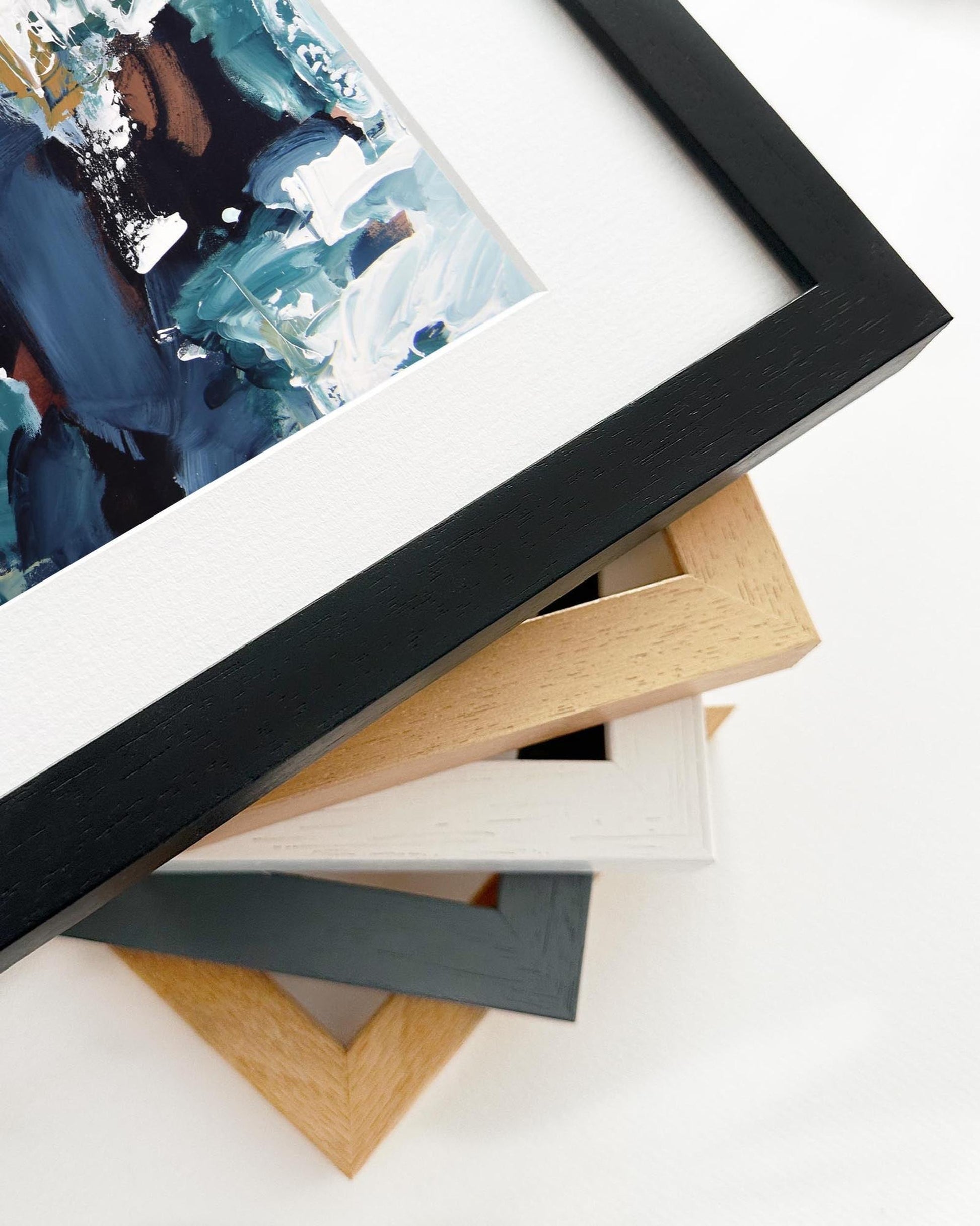 Stack of variously colored picture frames on a white surface with a framed abstract painting in the foreground.