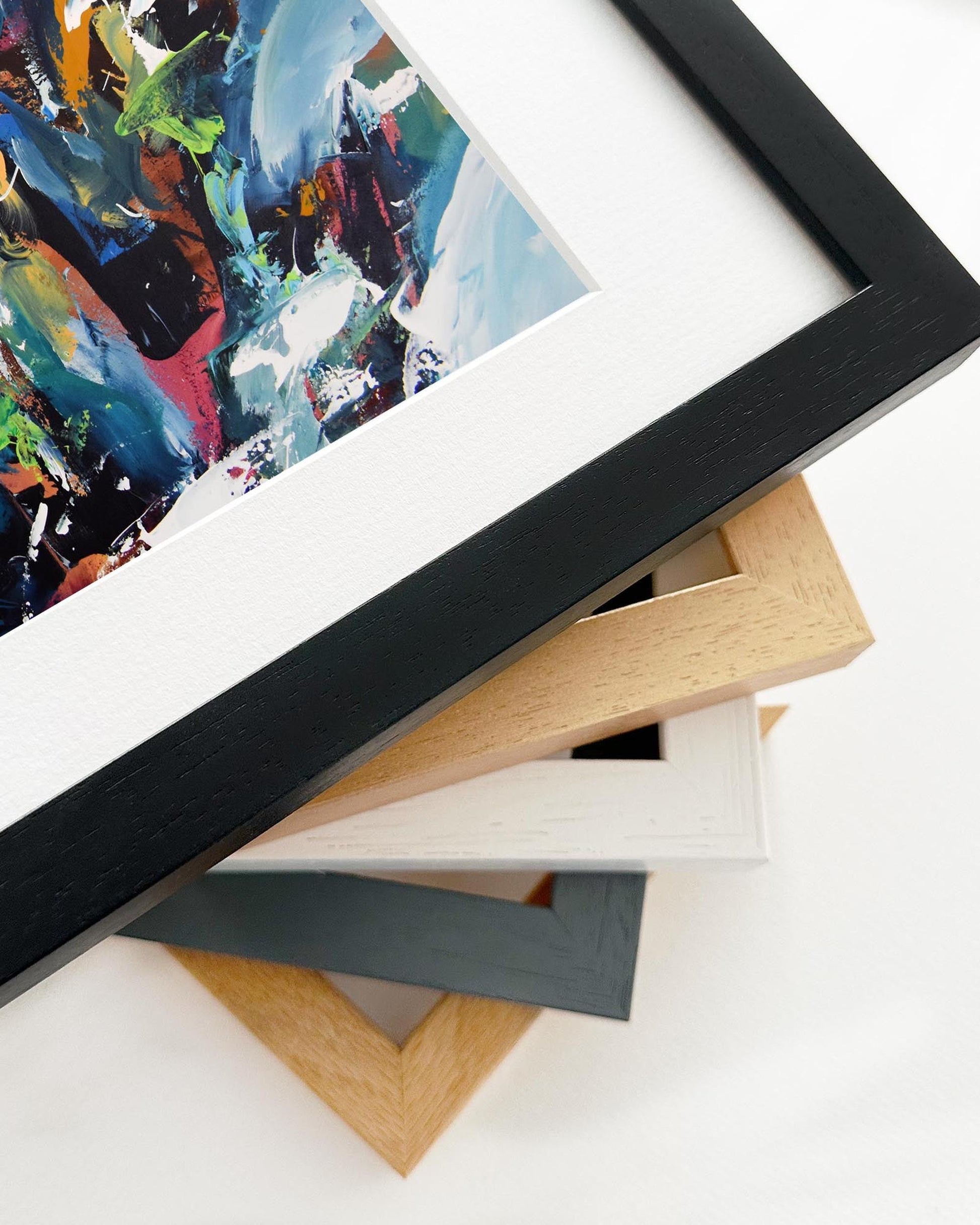 Stack of variously colored picture frames on a white surface with a colorful abstract painting in the background.