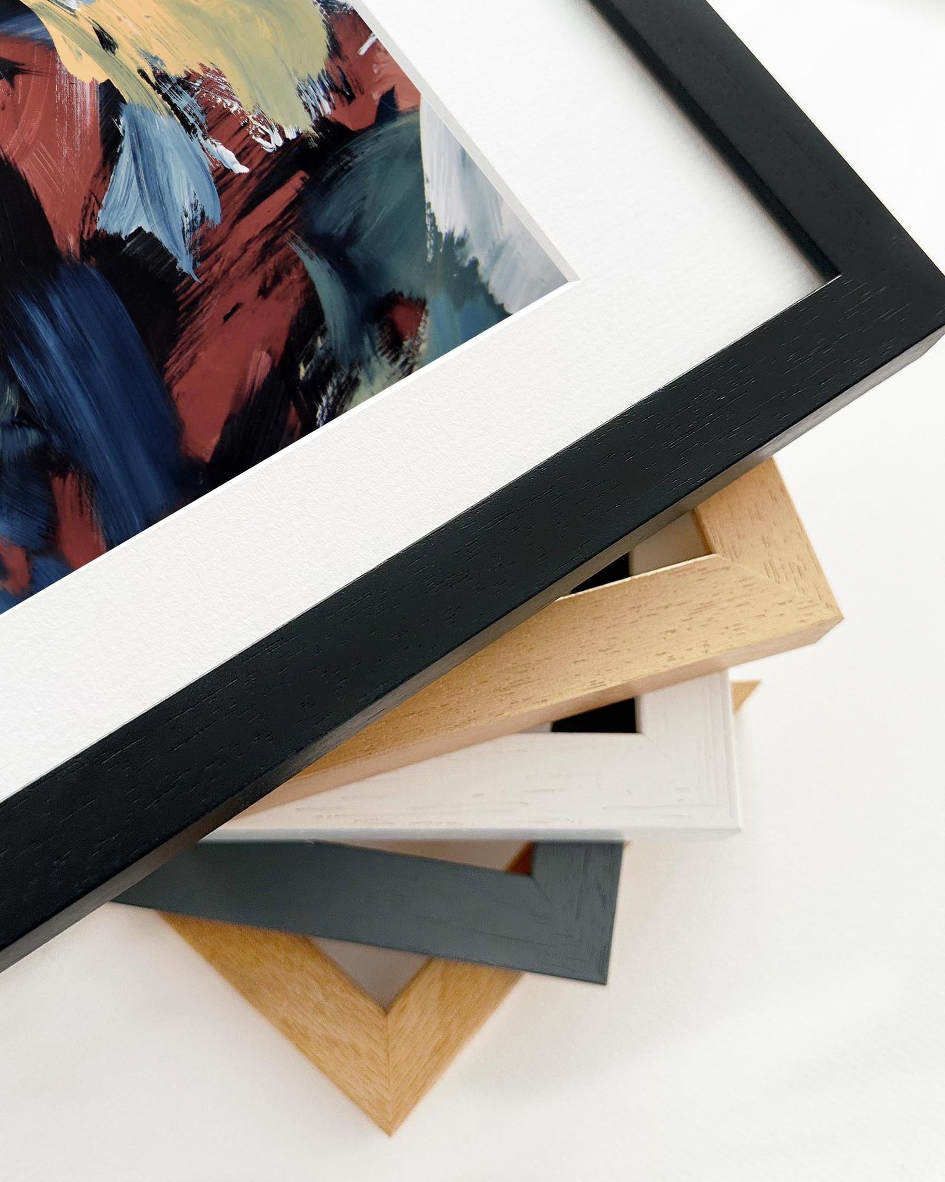 Stack of variously colored picture frames on a white surface with a partially visible abstract painting.