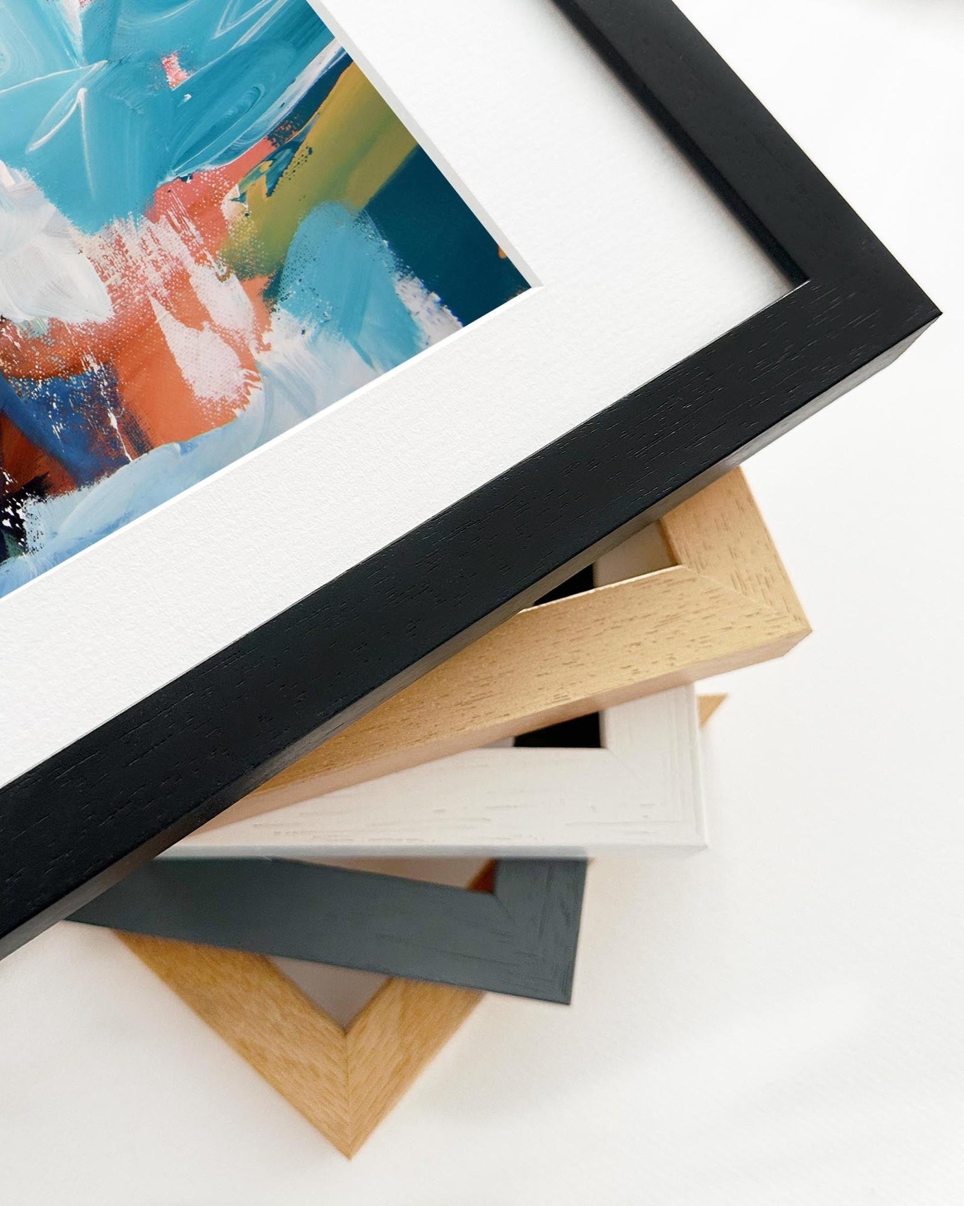 Stack of variously colored picture frames on a white surface with a partially visible abstract painting.
