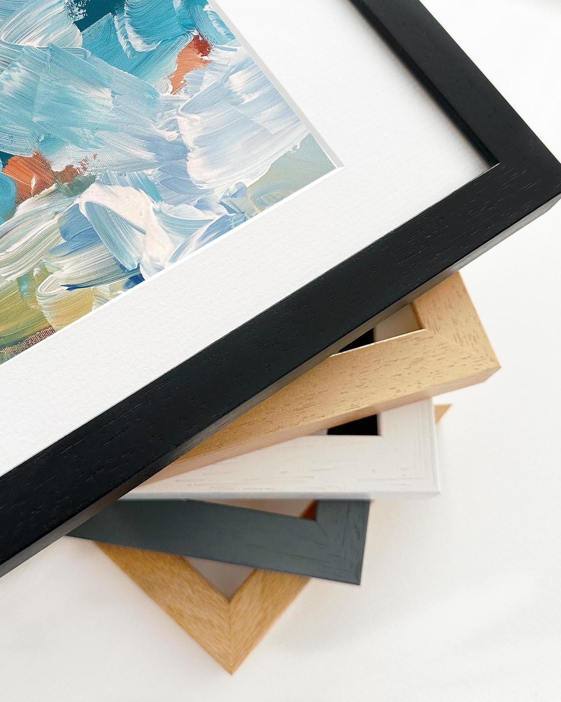Close-up of a black framed painting with additional frames stacked below on a white surface.