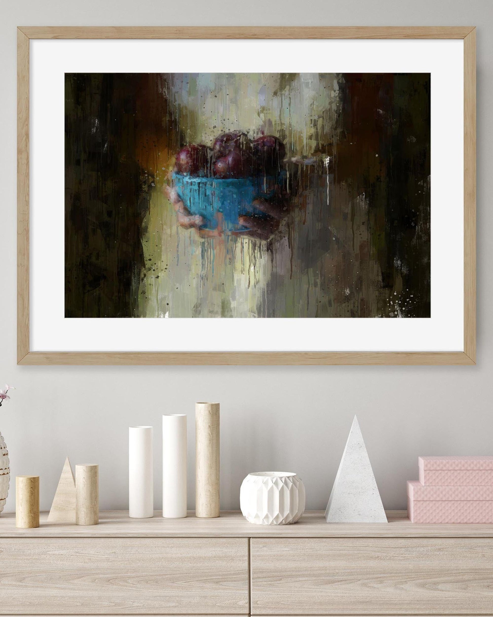 Framed abstract painting of a blue bowl with grapes on a wall above a wooden shelf with decorative items.
