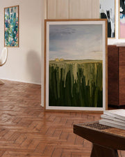 Framed painting of a landscape with green fields and a blue sky in a room with wooden flooring.
