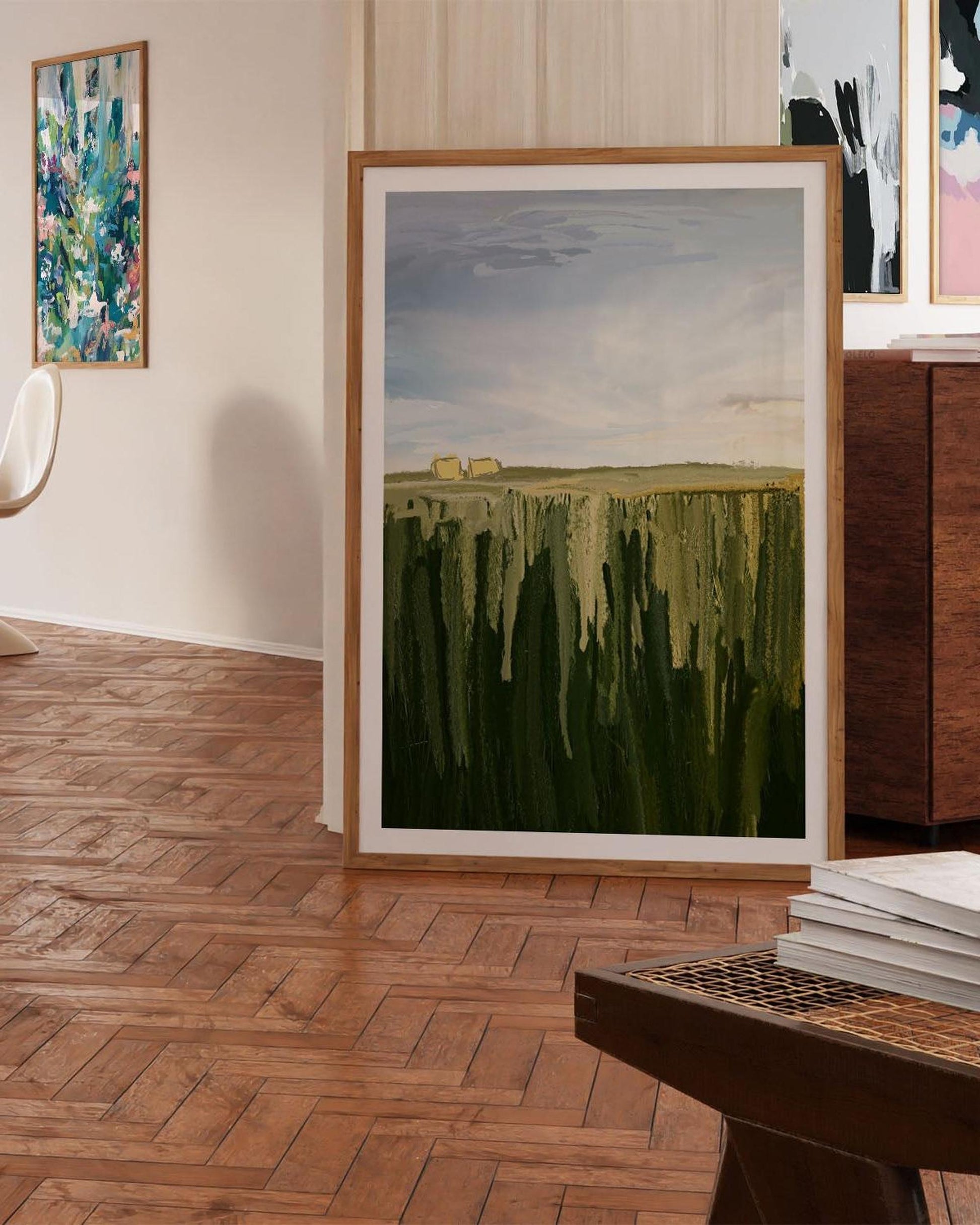 Framed painting of a landscape with green fields and a blue sky in a room with wooden flooring.