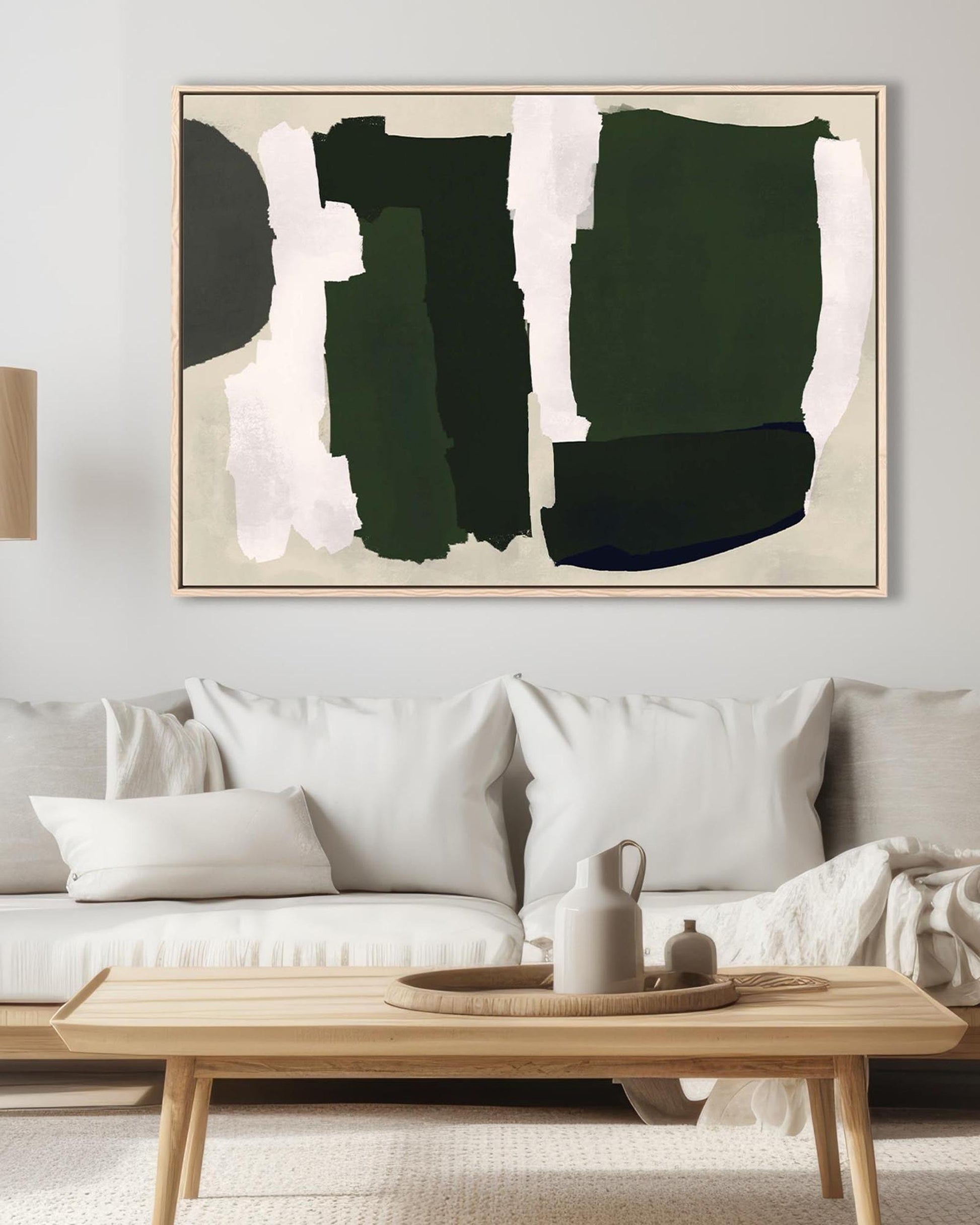 A canvas art print featuring abstract shapes in green and dark tones, displayed above a white sofa in a room with minimalist decor.