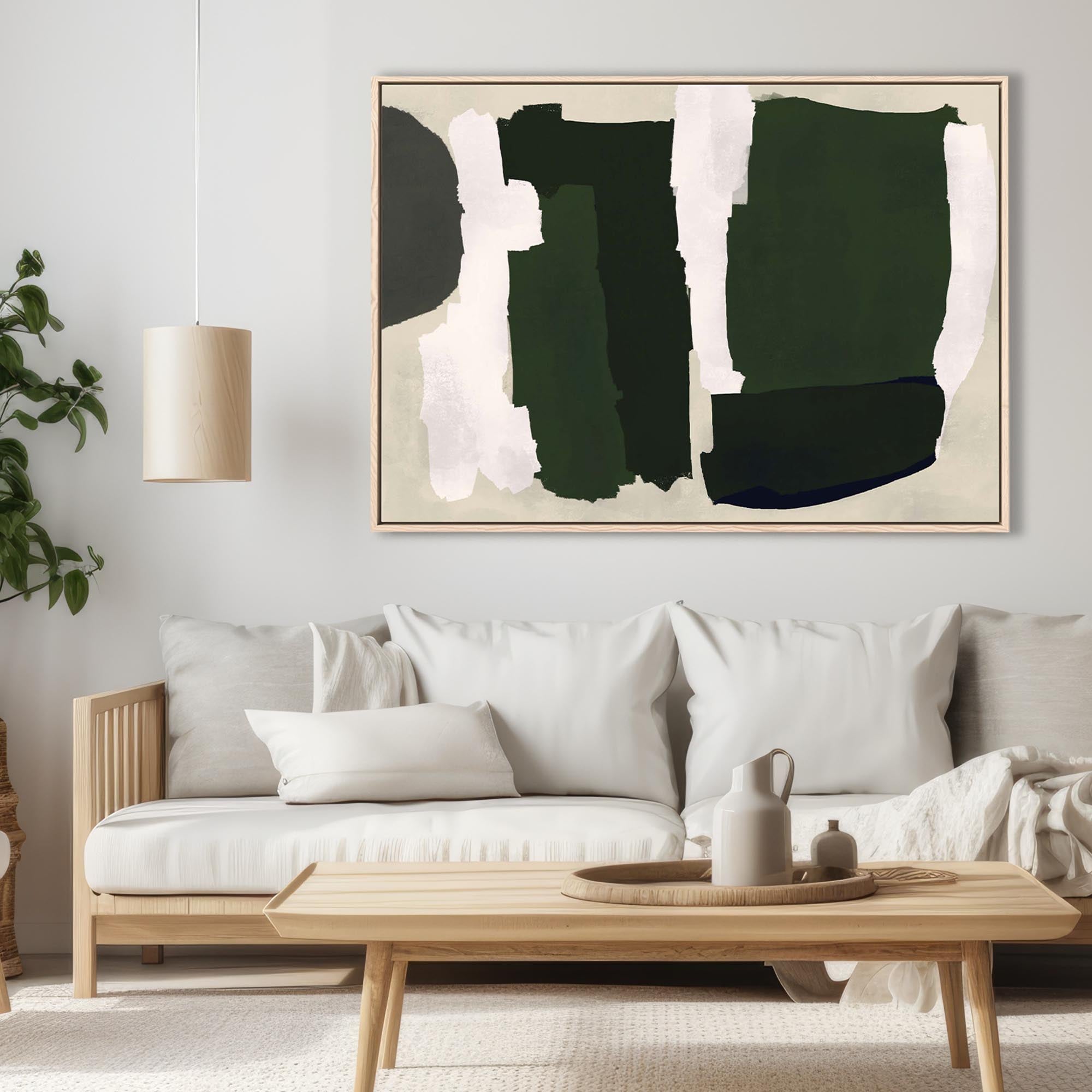 A canvas art print featuring abstract shapes in green and dark tones, displayed above a white sofa in a room with minimalist decor.