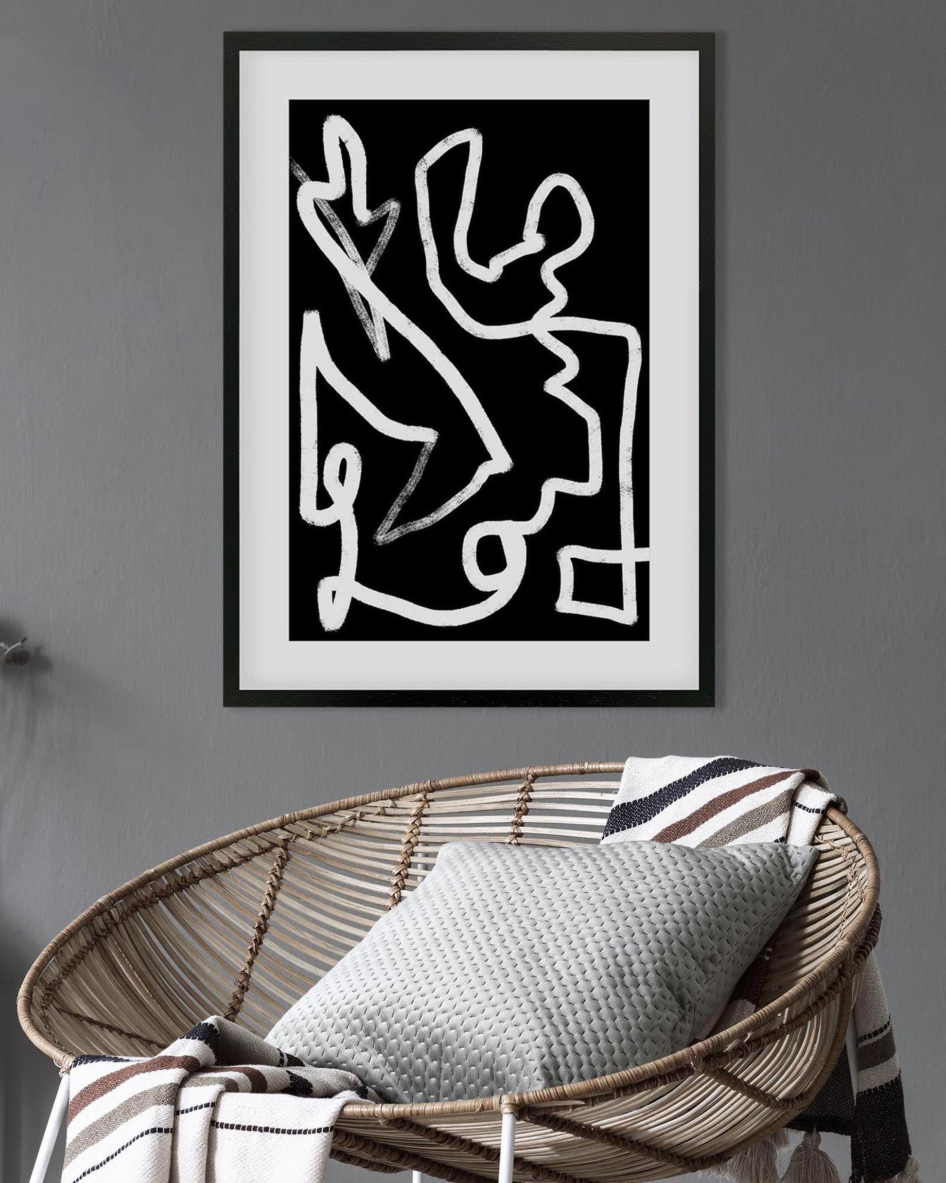 Framed abstract artwork on a gray wall above a wicker chair with a cushion.