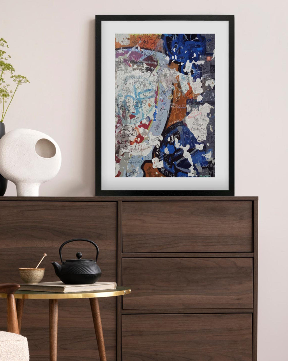 Framed abstract art piece on a wall above a wooden dresser with a teapot and small table.