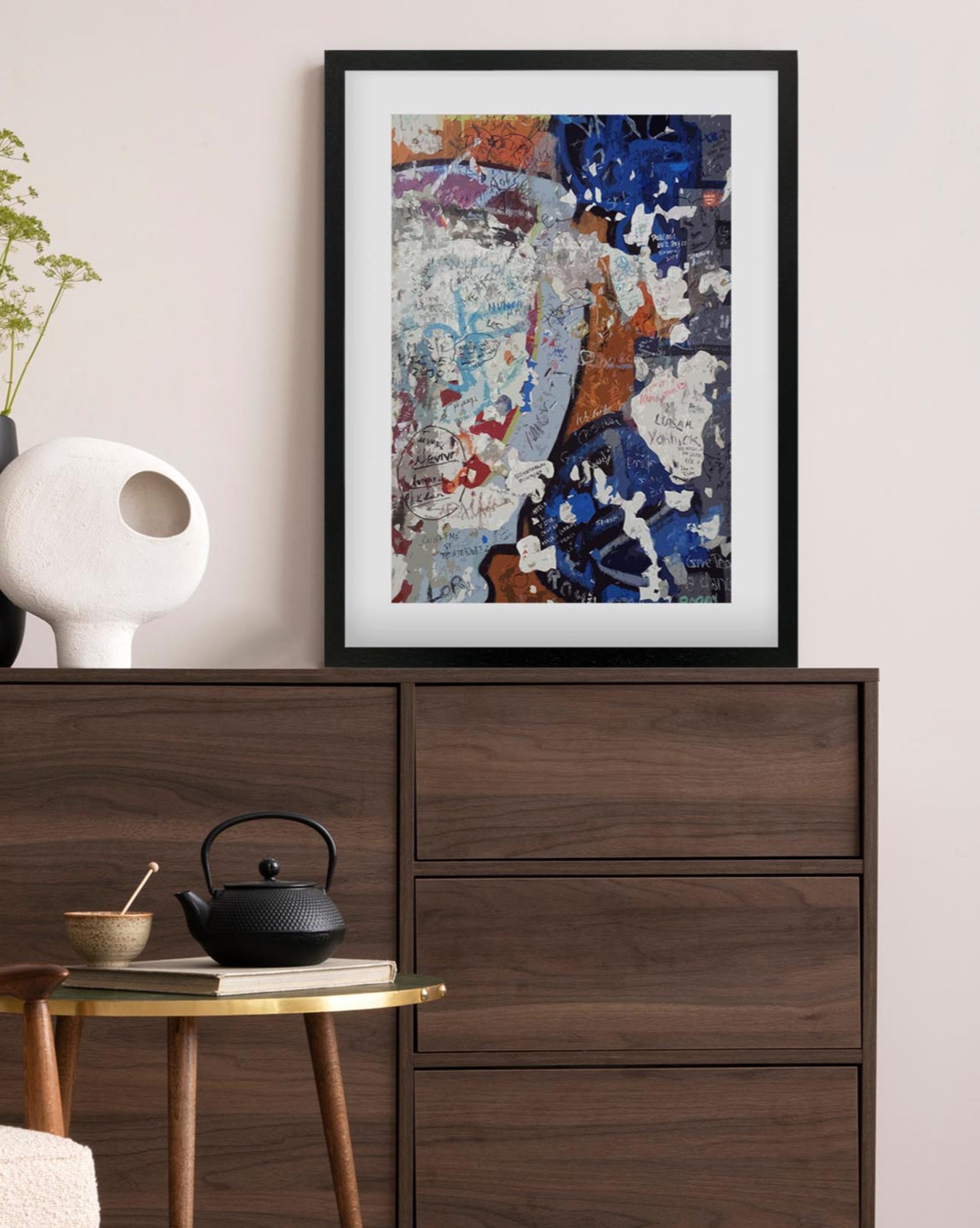 Framed abstract art piece on a wall above a wooden dresser with a teapot and small table.