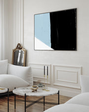 A framed canvas art print featuring bold abstract color blocks in black and blue, displayed above a white sofa in a bright living room setting.