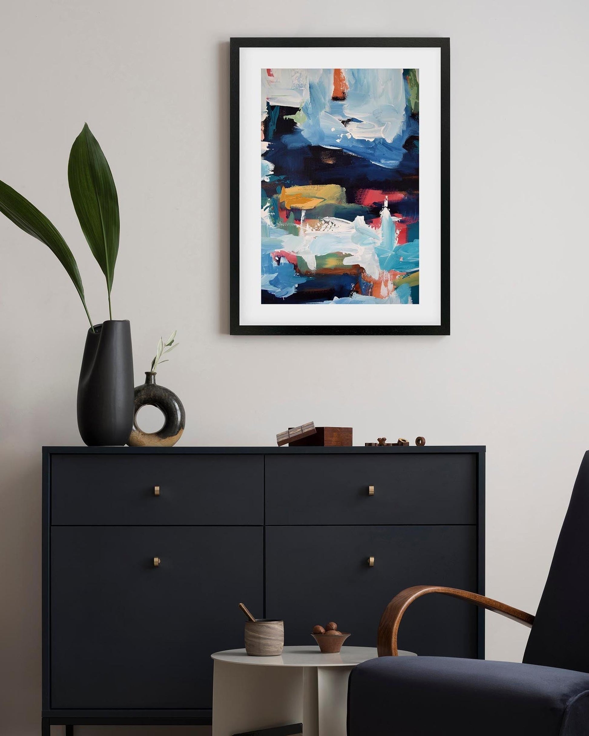 Framed abstract painting above a black dresser in a room with a plant and chair.