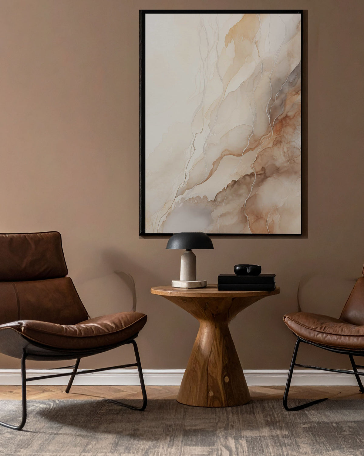A framed abstract wall art print with flowing beige, cream, and tan marble-like textures hangs on a warm taupe wall above a small wooden side table, flanked by two brown leather chairs in a styled interior.