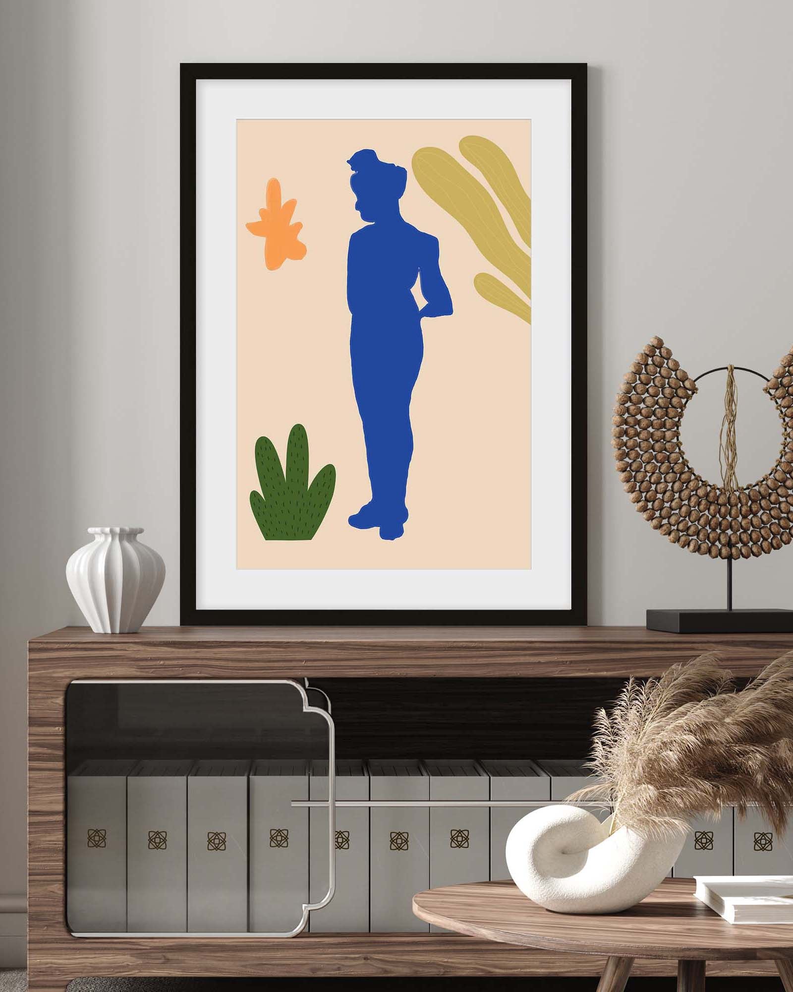 Framed abstract art print of a blue figure with green and yellow elements on a beige background, displayed on a wooden console table.