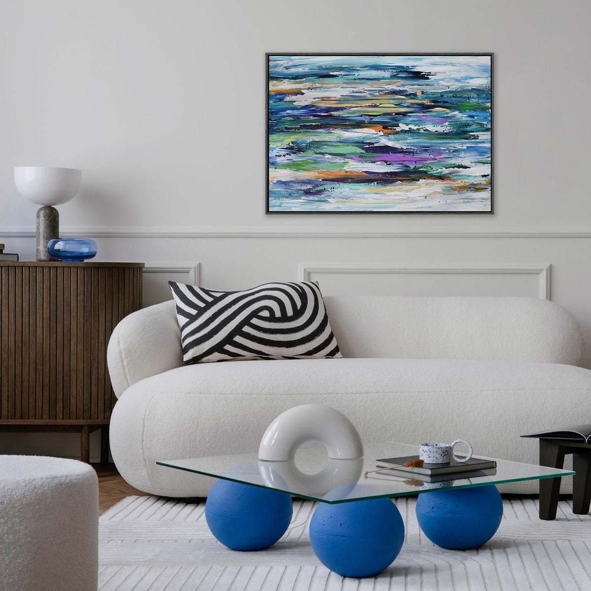 A framed abstract canvas painting with blue, green, purple, yellow, and white brushstrokes hangs on a white wall above a modern white sofa in a styled living room.