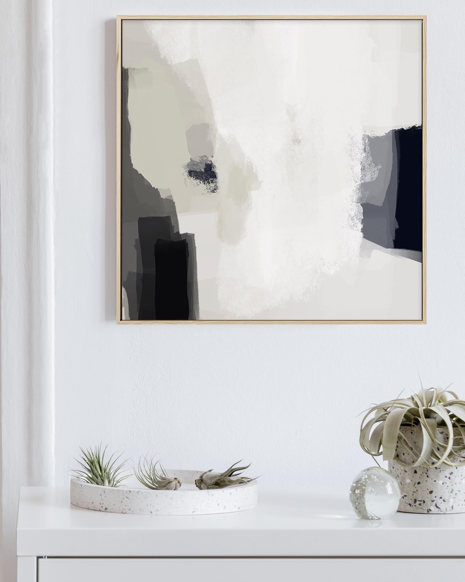 Abstract art piece in a gold frame on a white wall above a white console table with decorative plants.