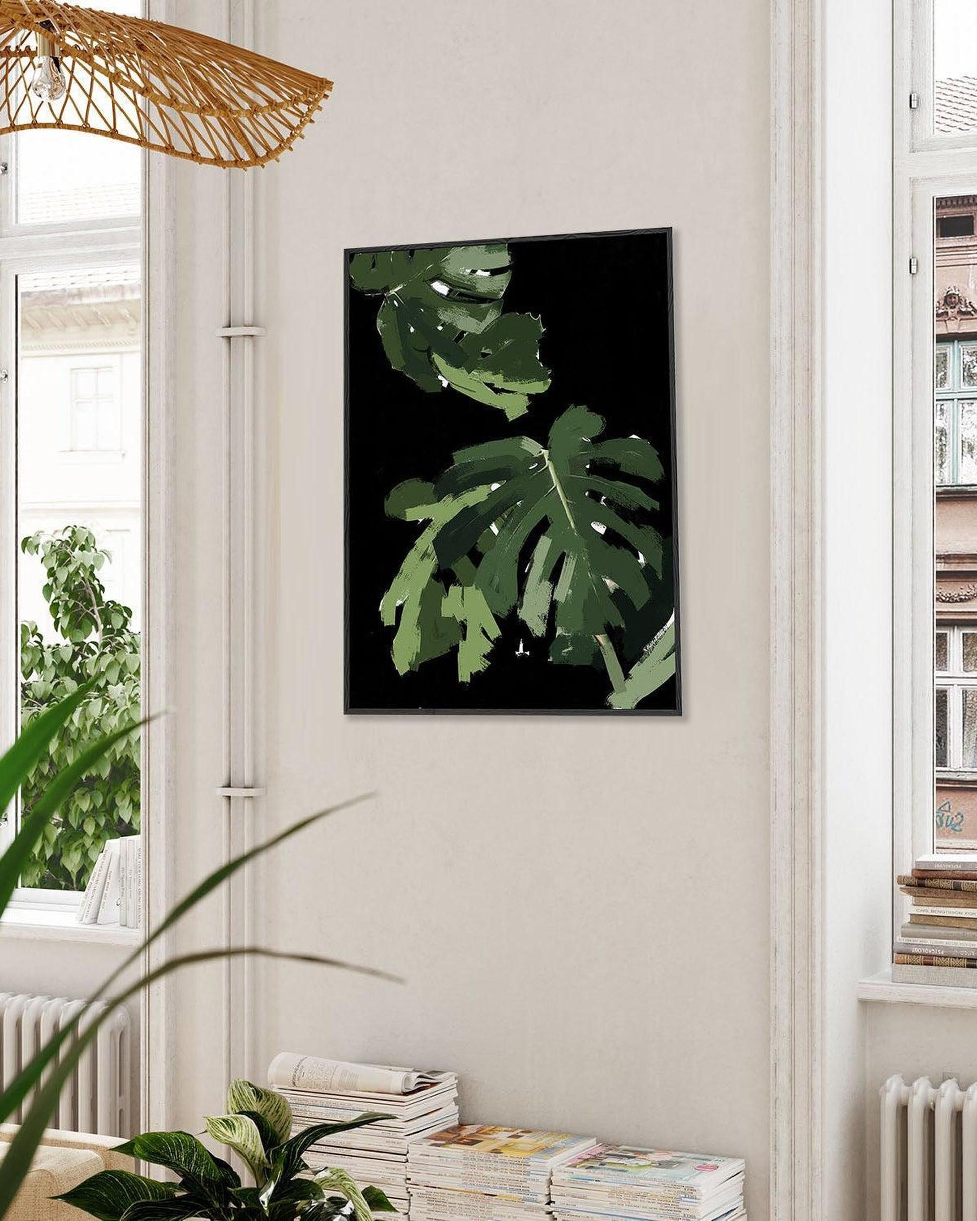 Framed artwork of green leaves on a wall in a room with plants and books.