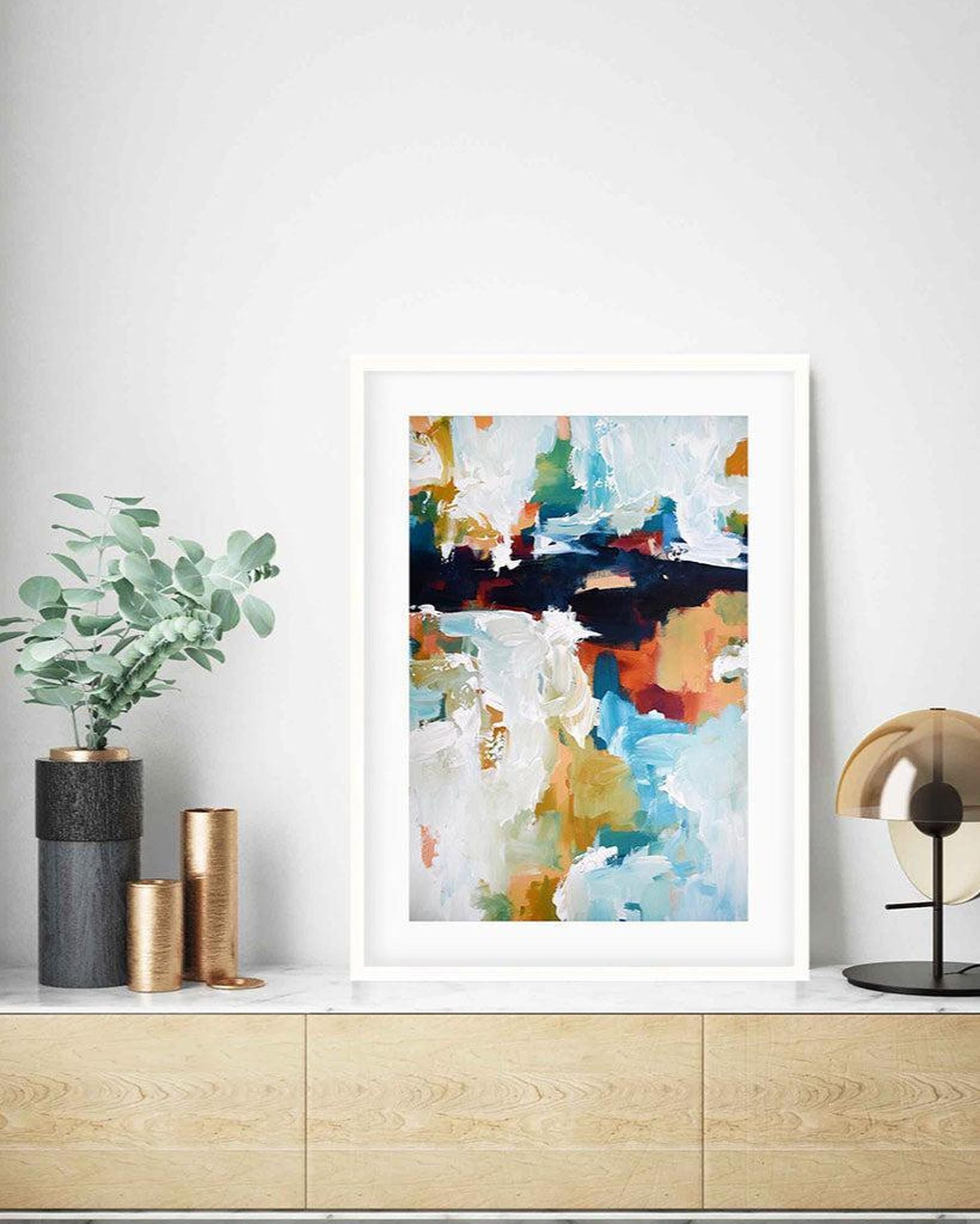 Framed abstract art print on a wall above a wooden cabinet with decorative items.