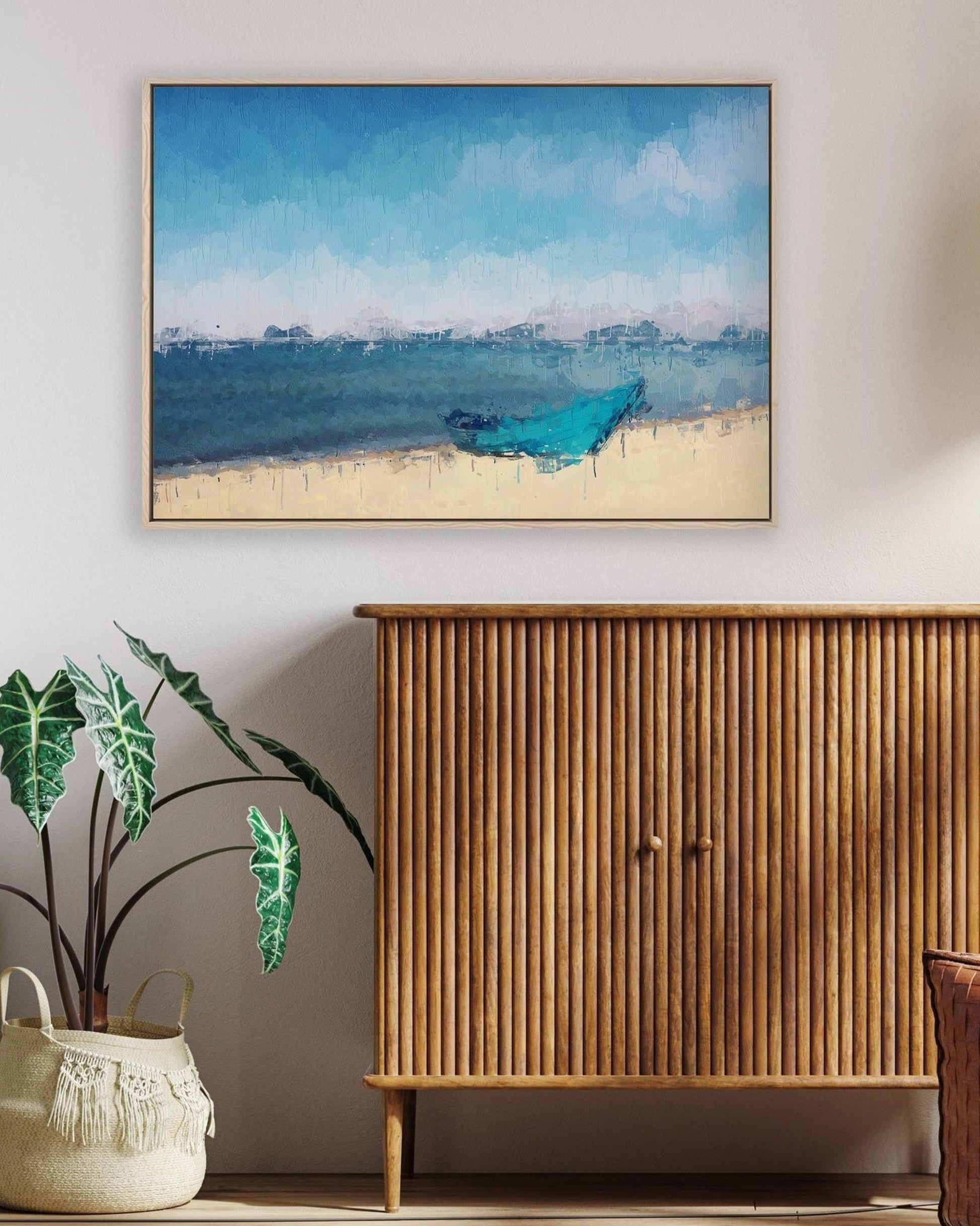 Framed artwork of a blue boat on a beach above a wooden cabinet with a plant and basket below.