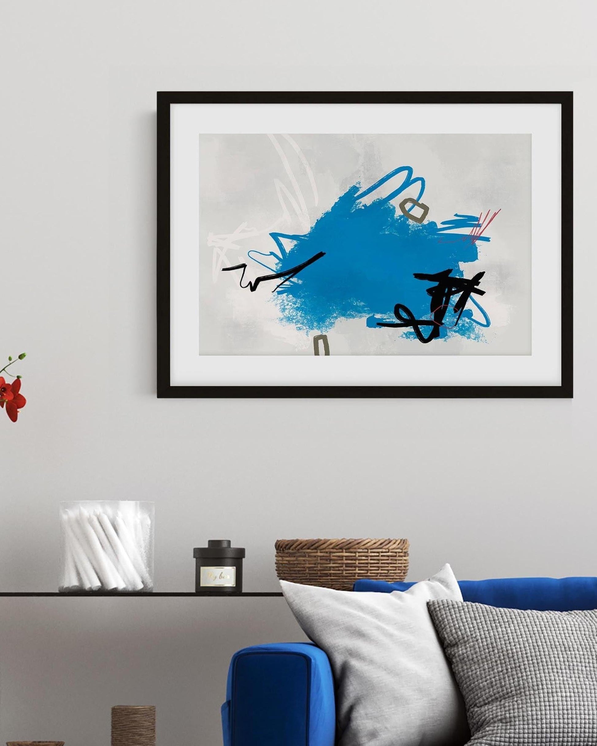 Abstract art piece with blue and black design in a black frame on a wall above a sofa.