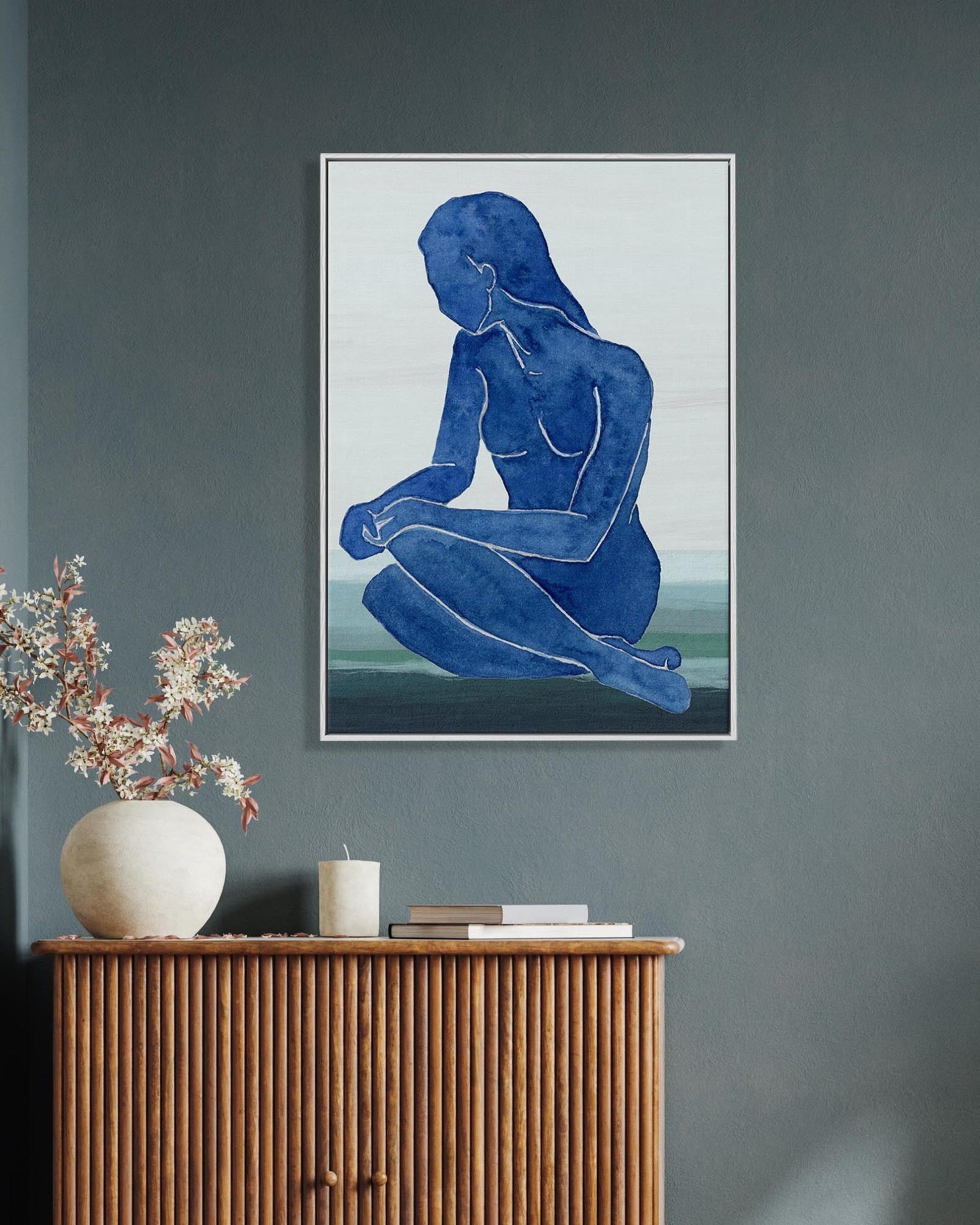 Framed artwork of a blue abstract figure on a gray wall above a wooden cabinet with decorative items.