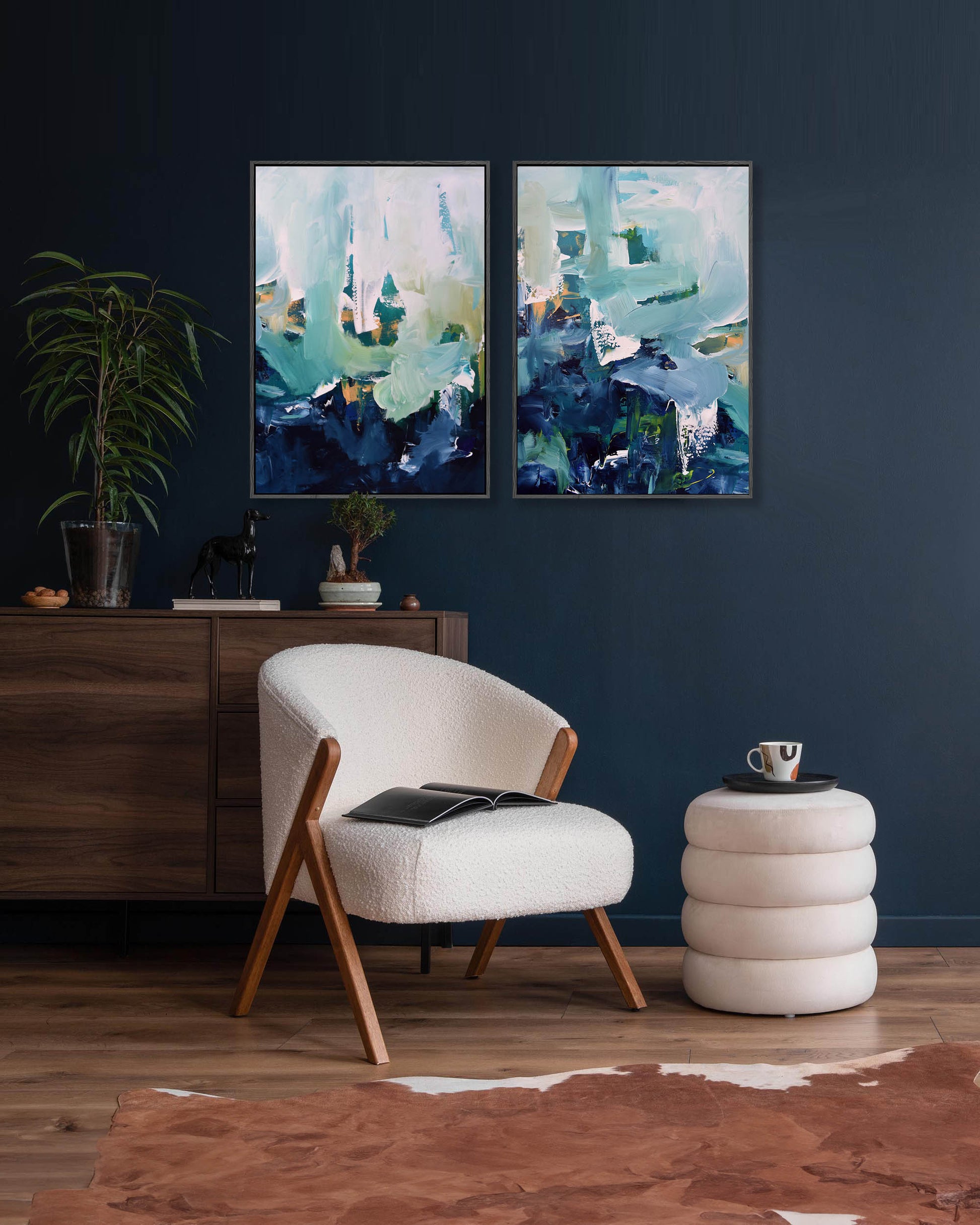 A set of two abstract canvas paintings featuring shades of blue and green, displayed above a modern chair and on a dark blue wall, with a plant and wooden cabinet in the background.