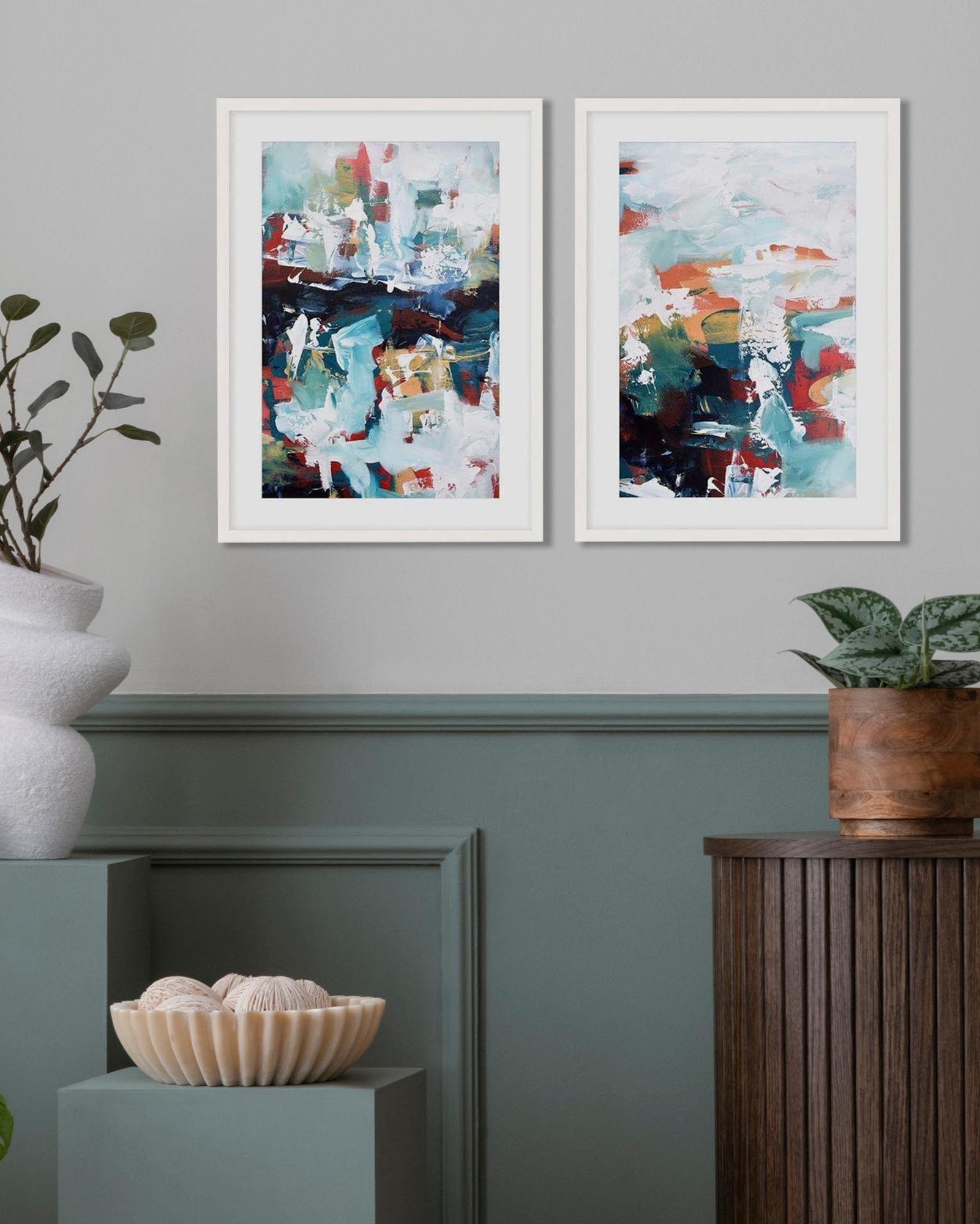 Two abstract paintings on a wall with decorative plants and vases below.