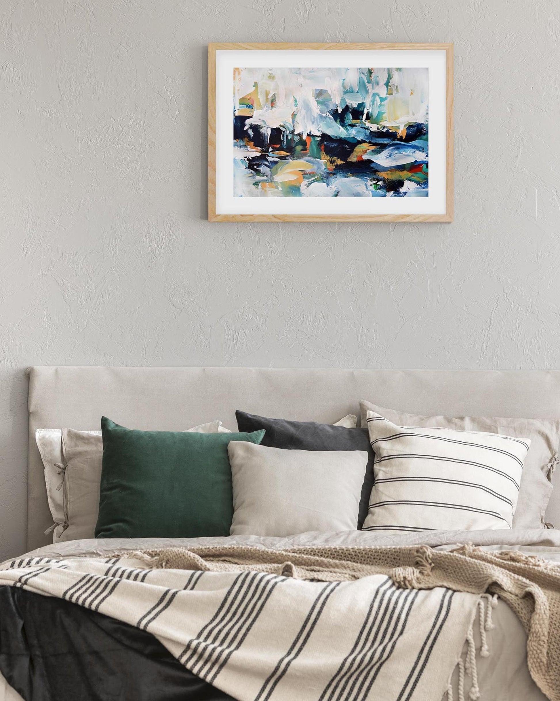Framed abstract painting on a wall above a bed with pillows and a blanket.