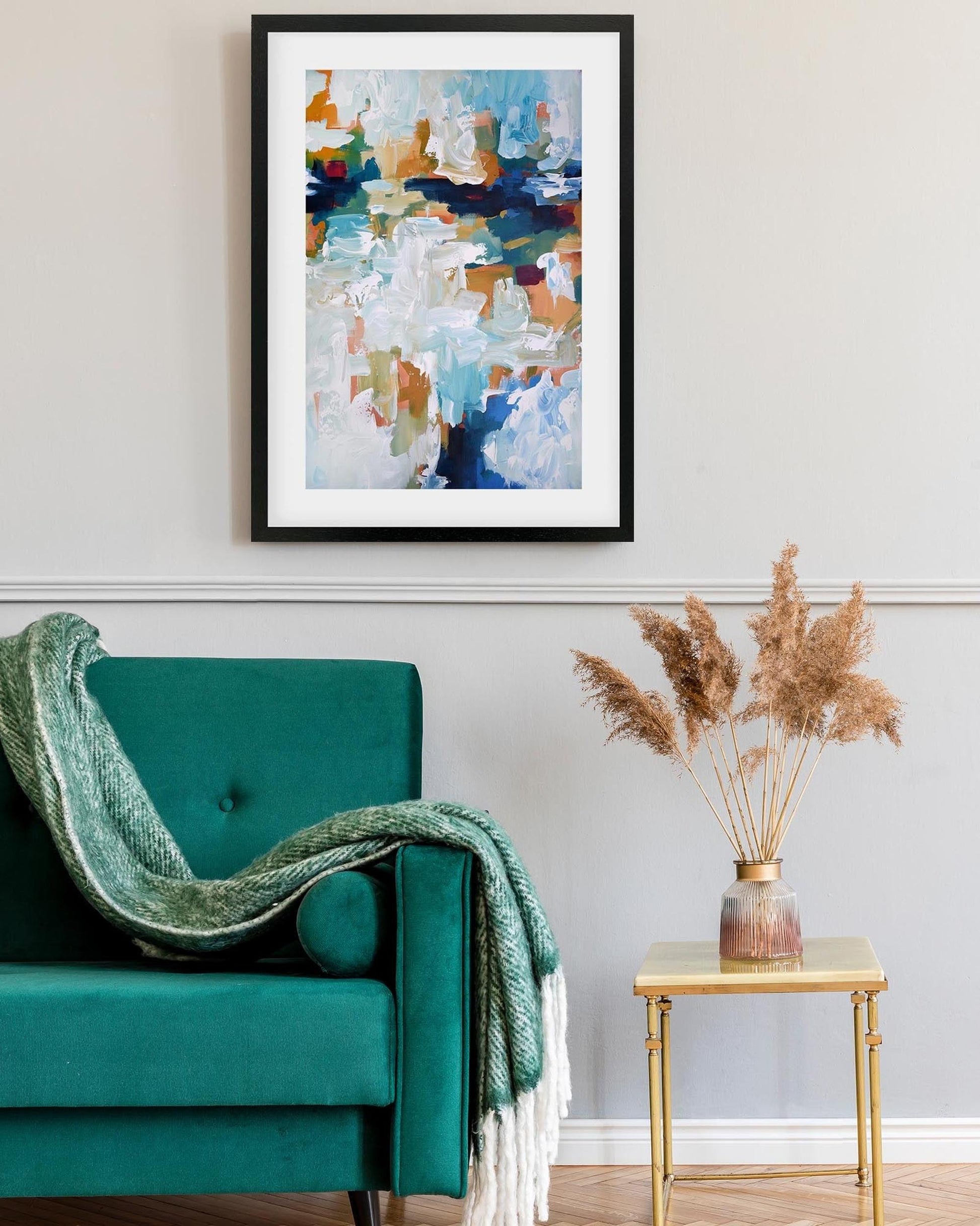 Teal armchair with a green blanket, gold side table with dried plants, and abstract art on the wall.
