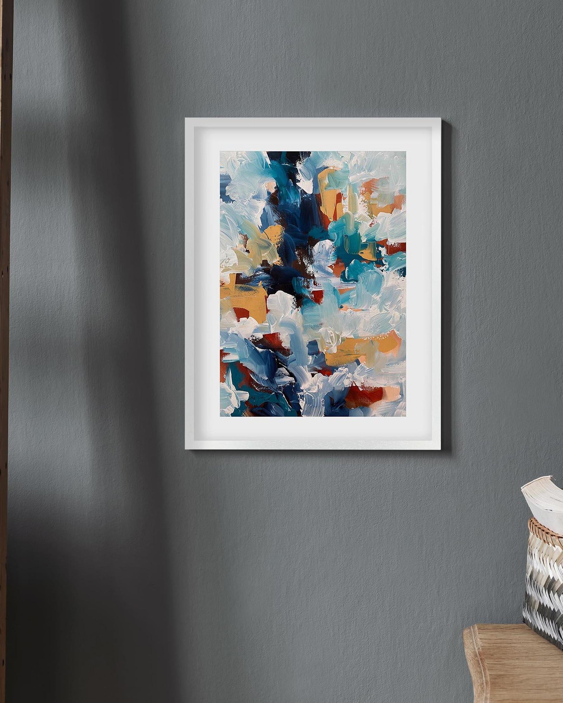 Framed abstract art print on a gray wall with a basket and cushion in the corner.