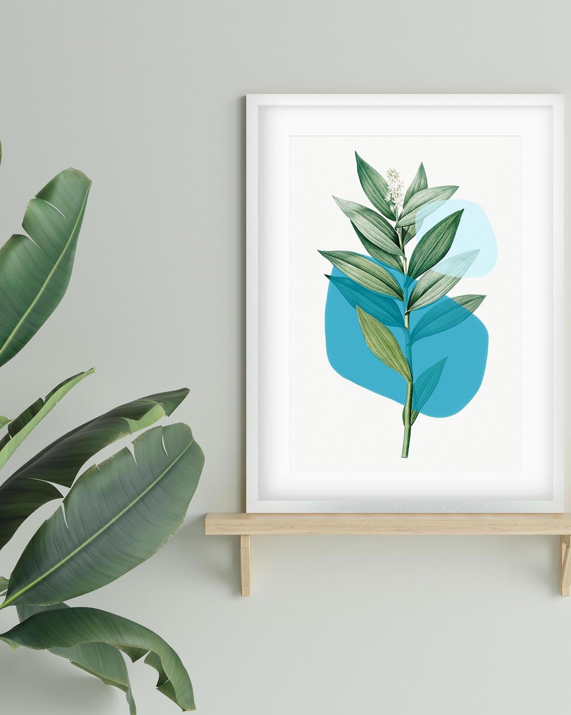 Framed botanical print with green leaves on a blue abstract background, displayed on a light gray wall.