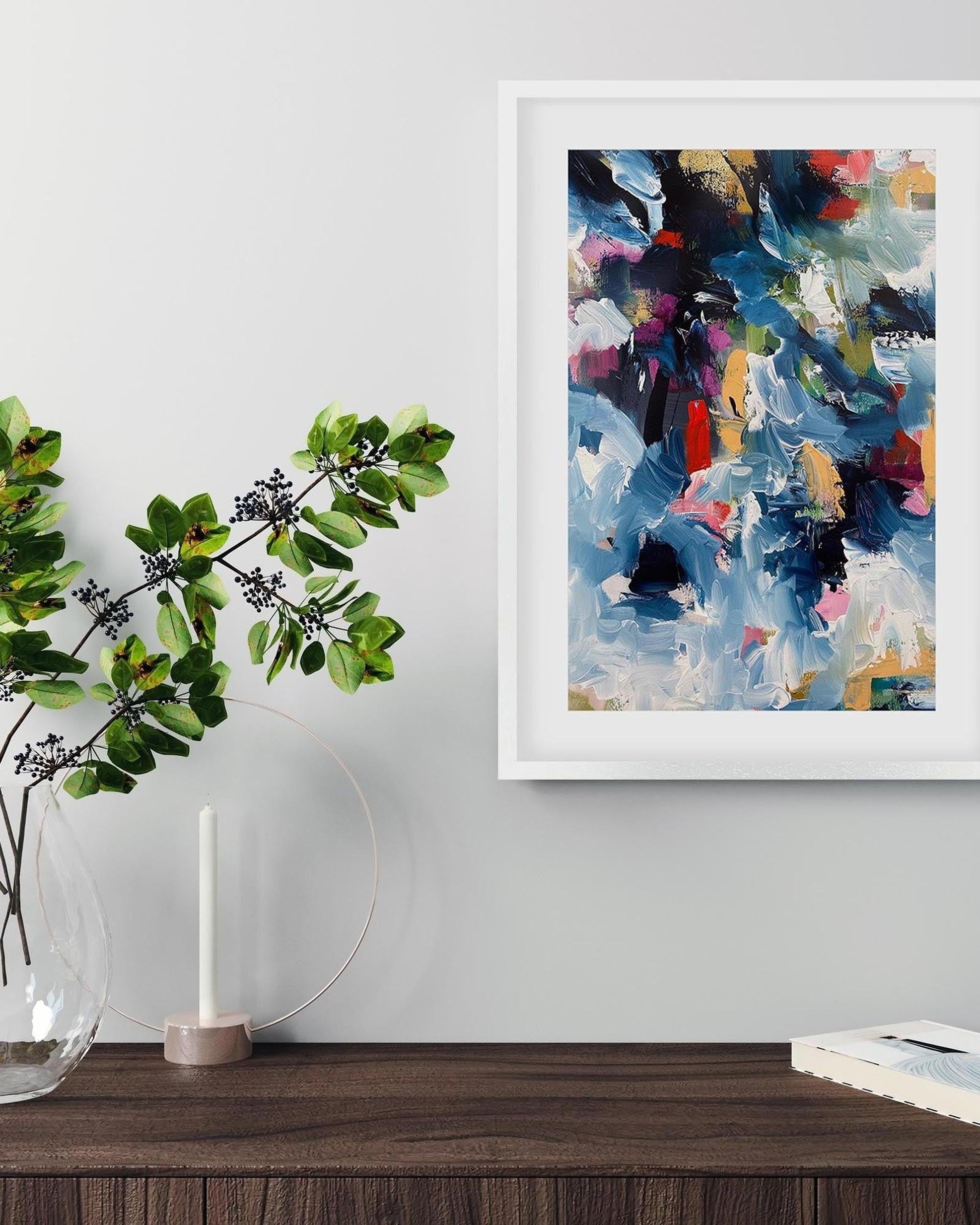 Abstract painting in a white frame on a wall above a wooden surface with a plant and books.