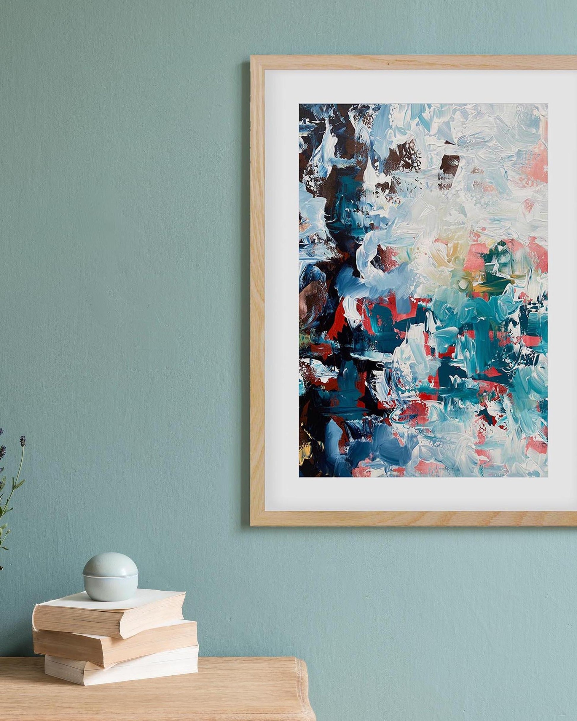 Framed abstract painting on a teal wall with books and a plant below.