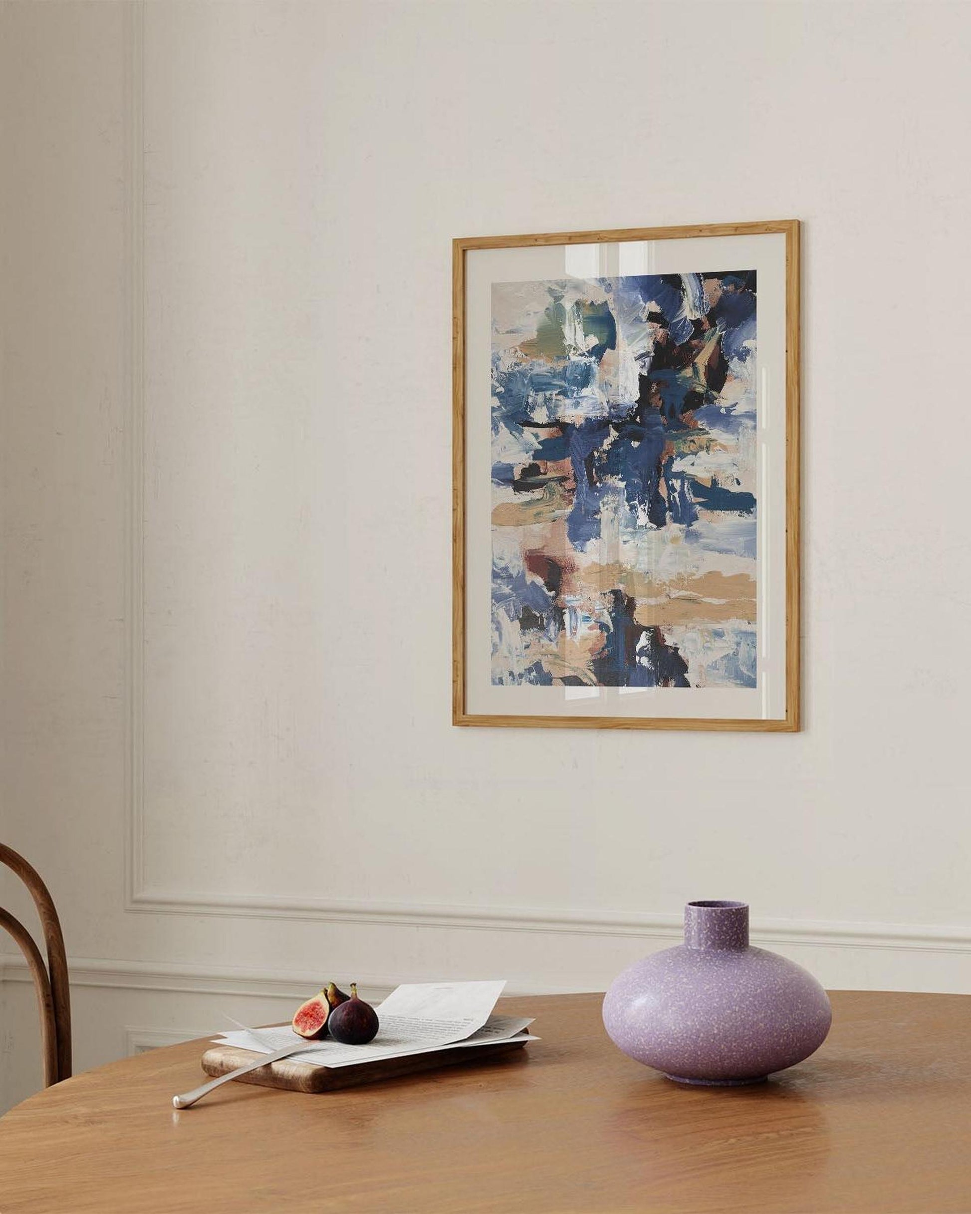 Abstract painting on a wall above a wooden table with a vase and fruit.