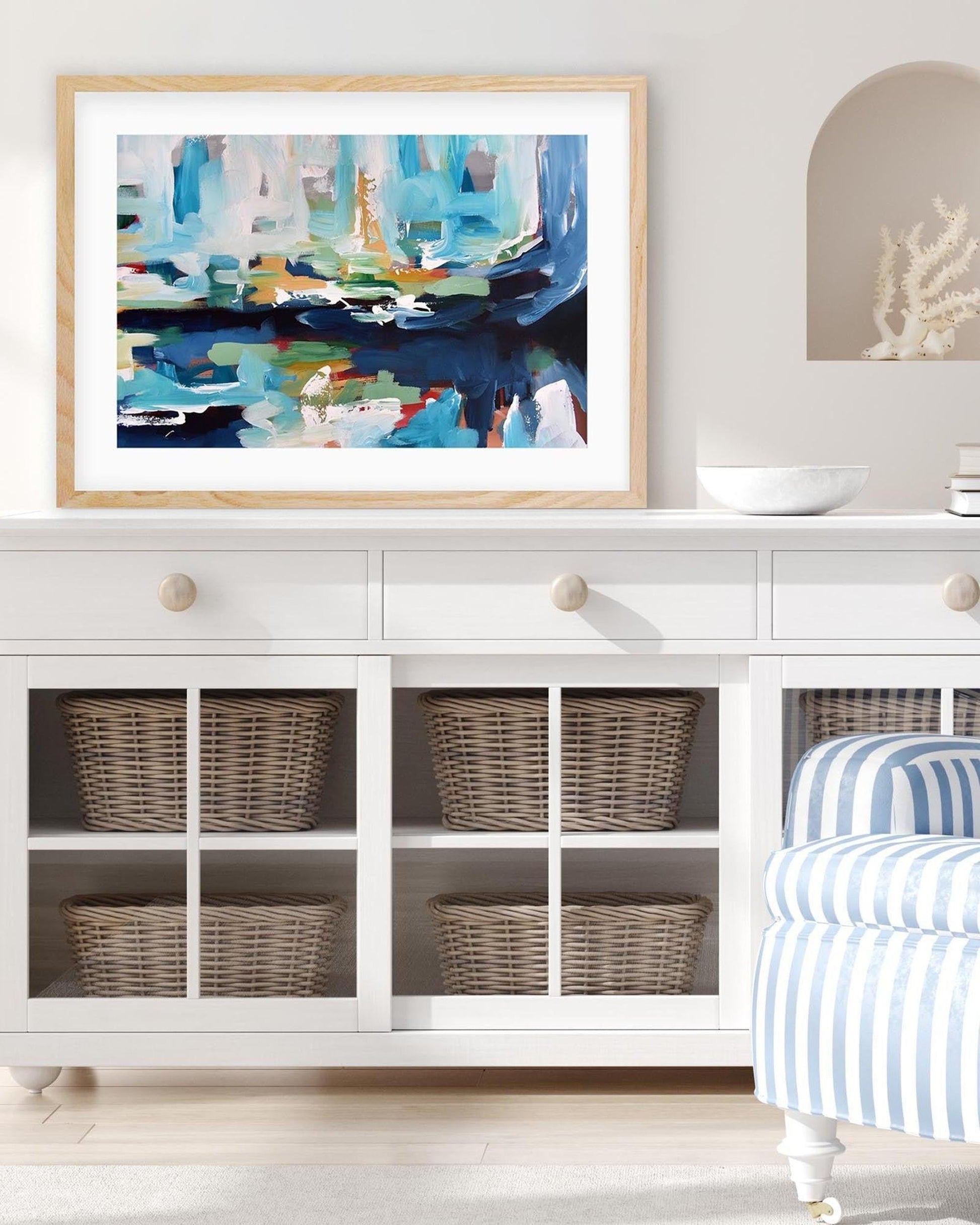 Framed abstract art above a white cabinet with wicker baskets, striped chair, and decorative items.