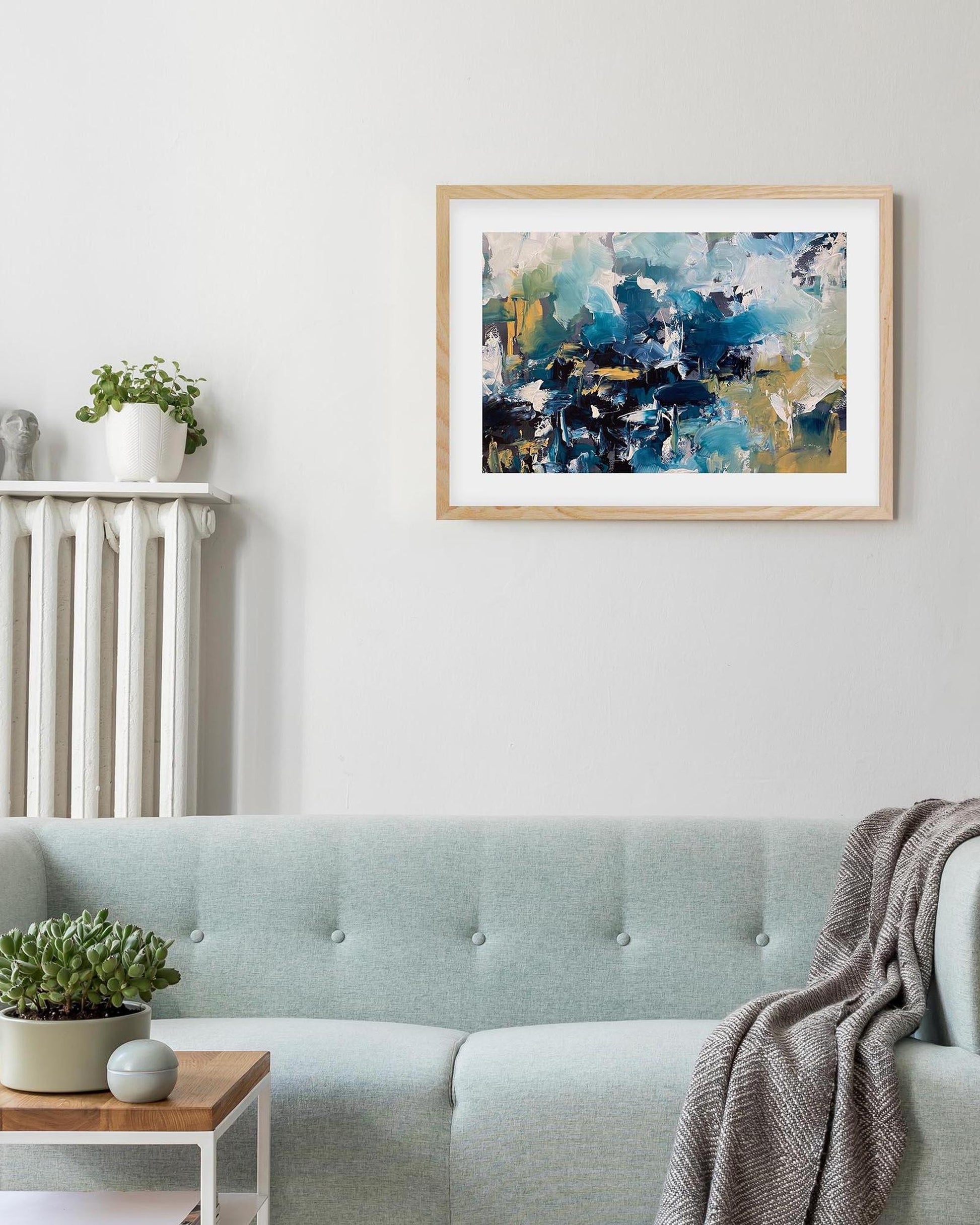 Abstract painting in a wooden frame on a wall above a light blue sofa with decorative elements.