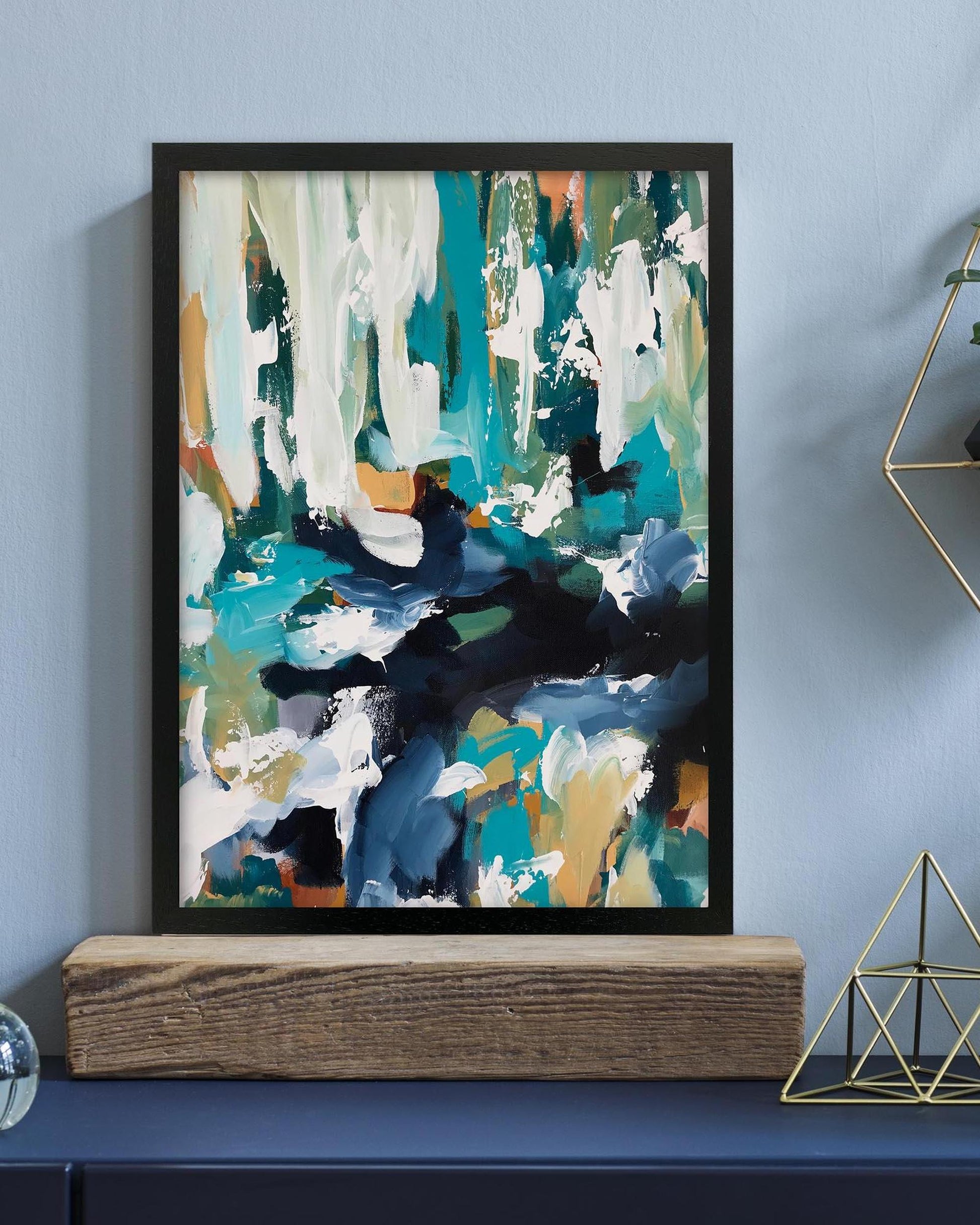 Abstract art print in a black frame on a blue wall above a wooden shelf.
