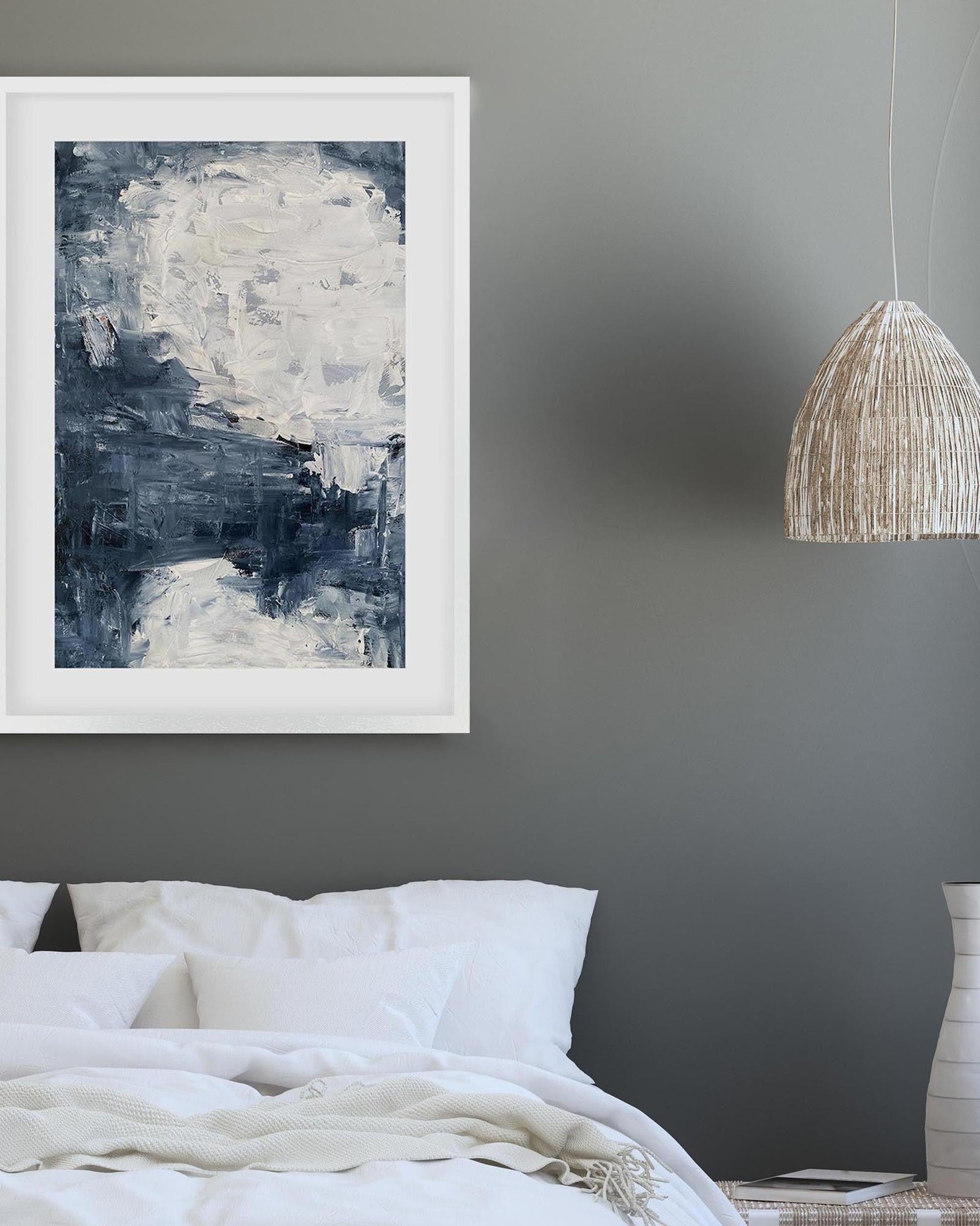 Abstract painting in a white frame on a gray wall above a bed.