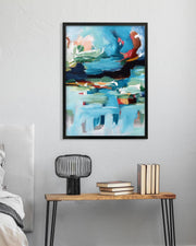 Framed abstract painting on a wall above a wooden side table with books and a lamp.